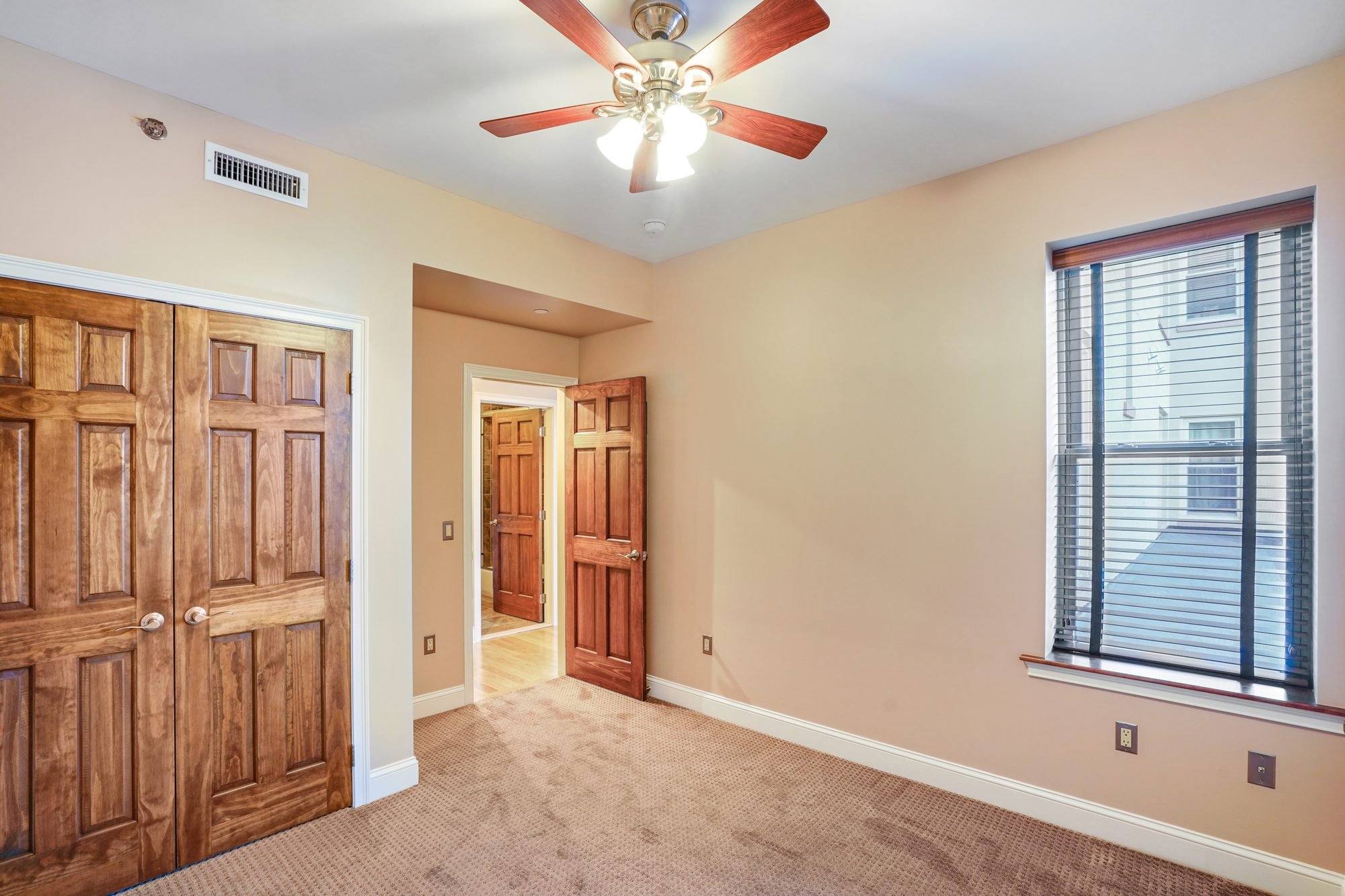Interior view of a residential room with beige walls, a window with blinds, a ceiling fan with wooden blades and lights, an open wooden closet door, and a door leading to another room or hallway.