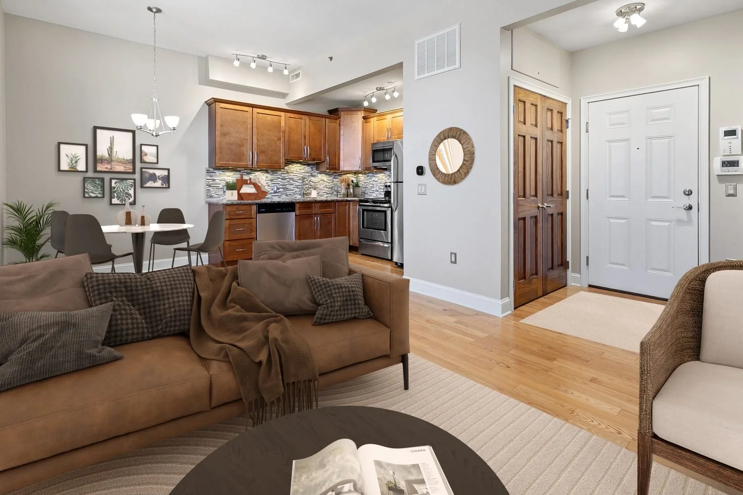 Living room with beige couch, pillows, blanket, and coffee table with open magazine; open kitchen with wooden cabinets, stainless steel appliances, and a dining area with a round table and four chairs; hardwood floors and decorative wall art; entry a
