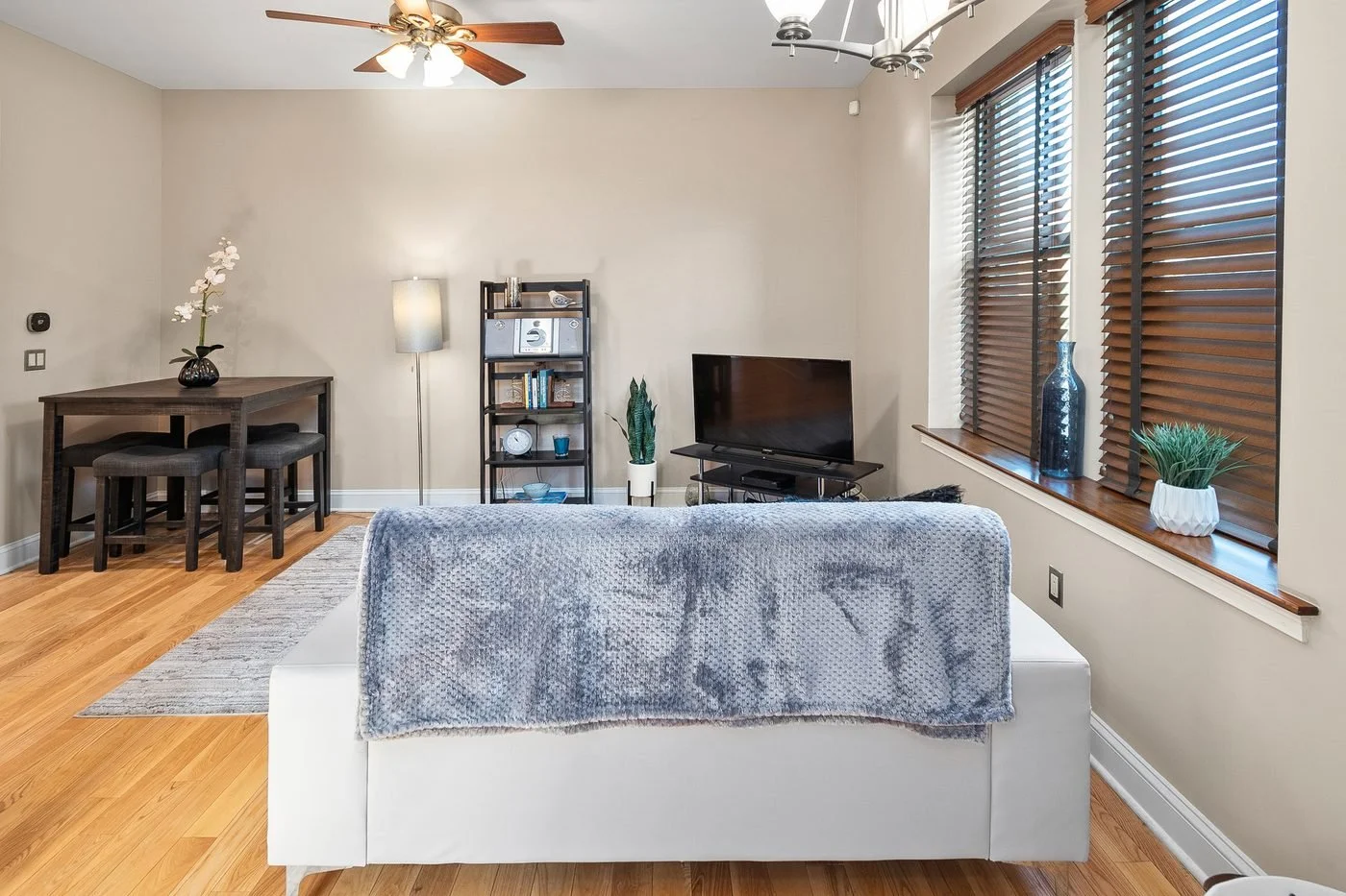 Living room with beige walls, wooden floors, a white sofa with a gray blanket, a TV on a black stand, a small dining table with two dark chairs, a tall window with wooden blinds, and various decorative items including plants, vases, and books.