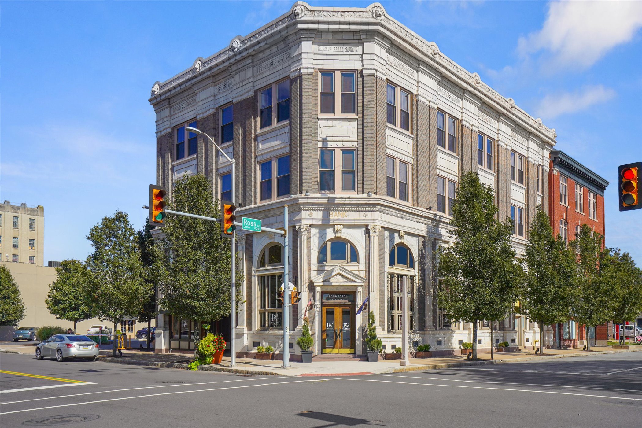 A multi-story historic brick building with ornate white stone detailing at the top, located on a city street corner. There are traffic lights and street signs visible, with cars parked along the street and trees lining the sidewalk.