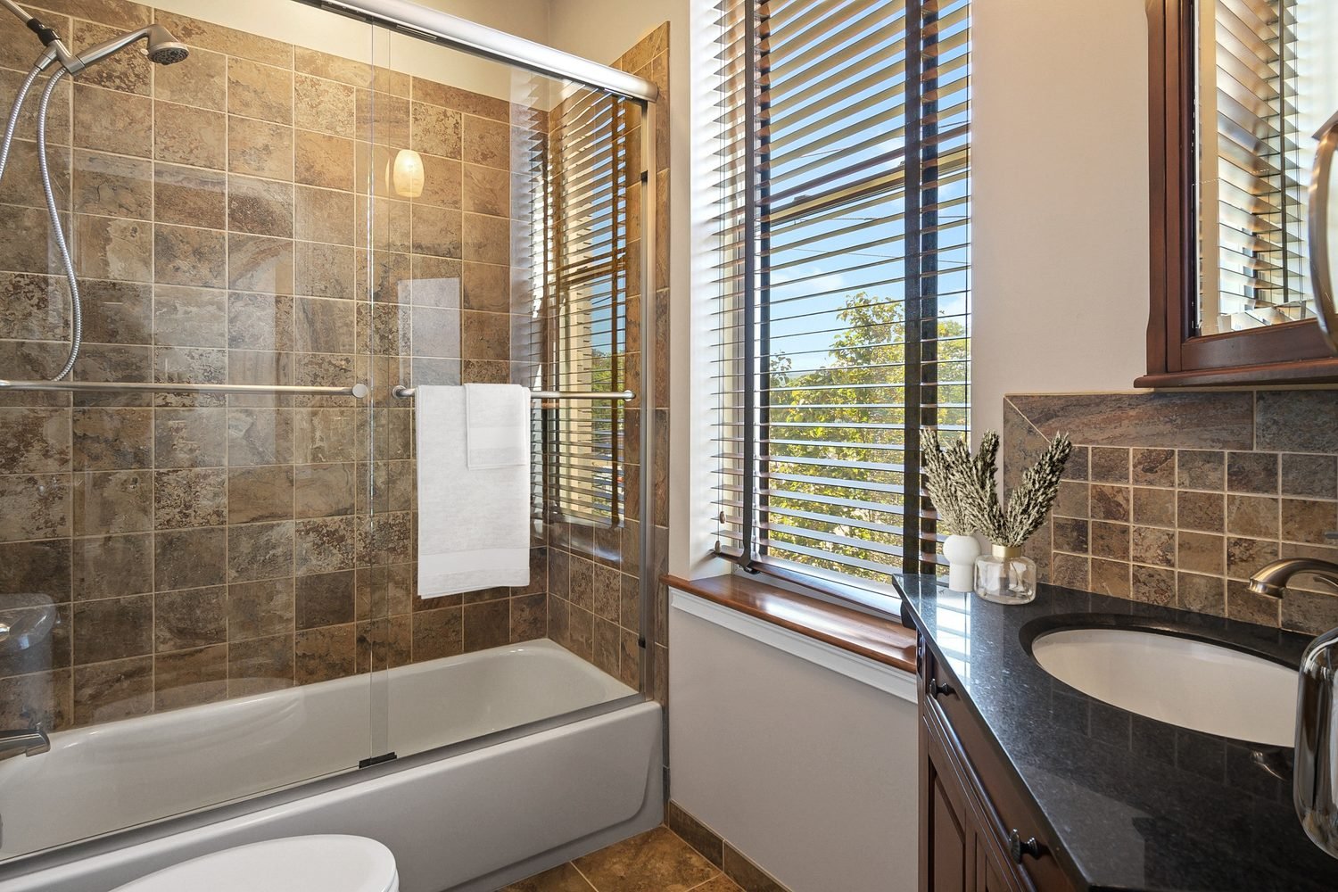 Bathroom with a bathtub, glass shower enclosure, brown tiled walls, and a large window with blinds