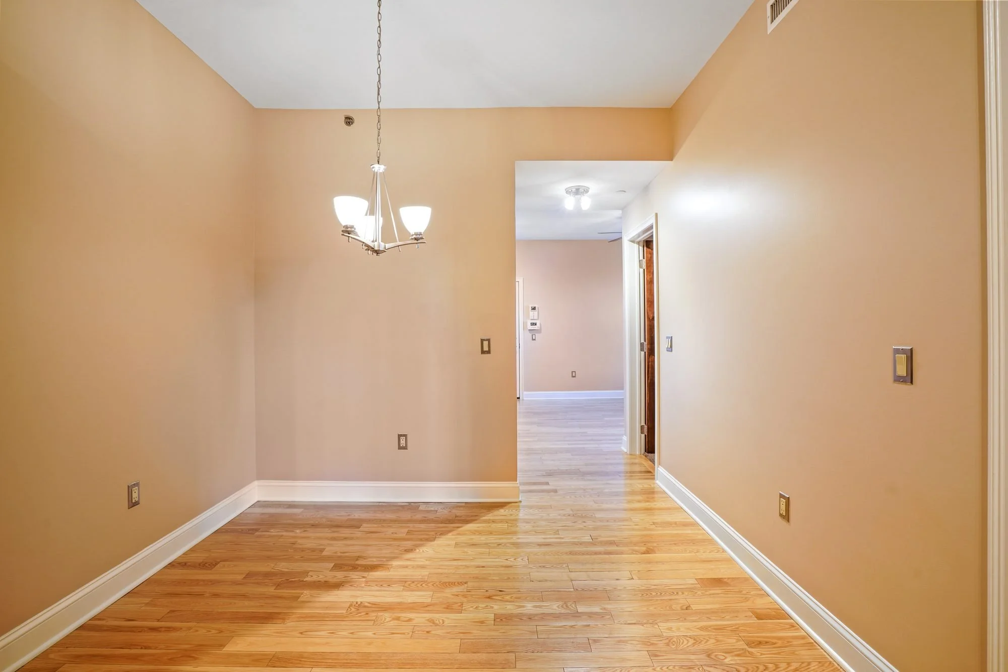Empty room with beige walls, hardwood floors, a chandelier, and a doorway leading to another room with ceiling lights.