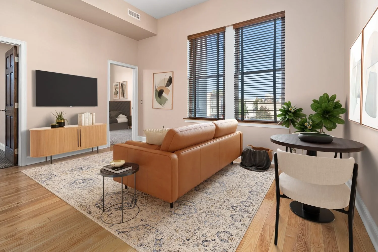 Living room with a tan leather sofa, wooden side table, and a patterned rug. TV mounted on the wall, with a wooden console below. Large windows with wooden blinds, green plants, and abstract artwork on the walls. Doorways leading to bedrooms.