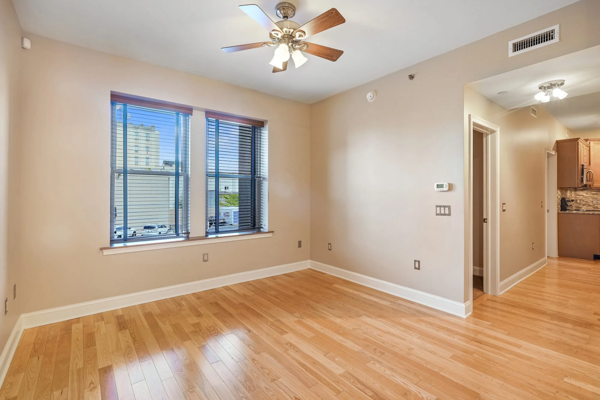 Living room with light-colored walls, wooden flooring, two large windows with blinds, ceiling fan with lights, thermostat on the wall, view of kitchen with wooden cabinets and backsplash through doorway.
