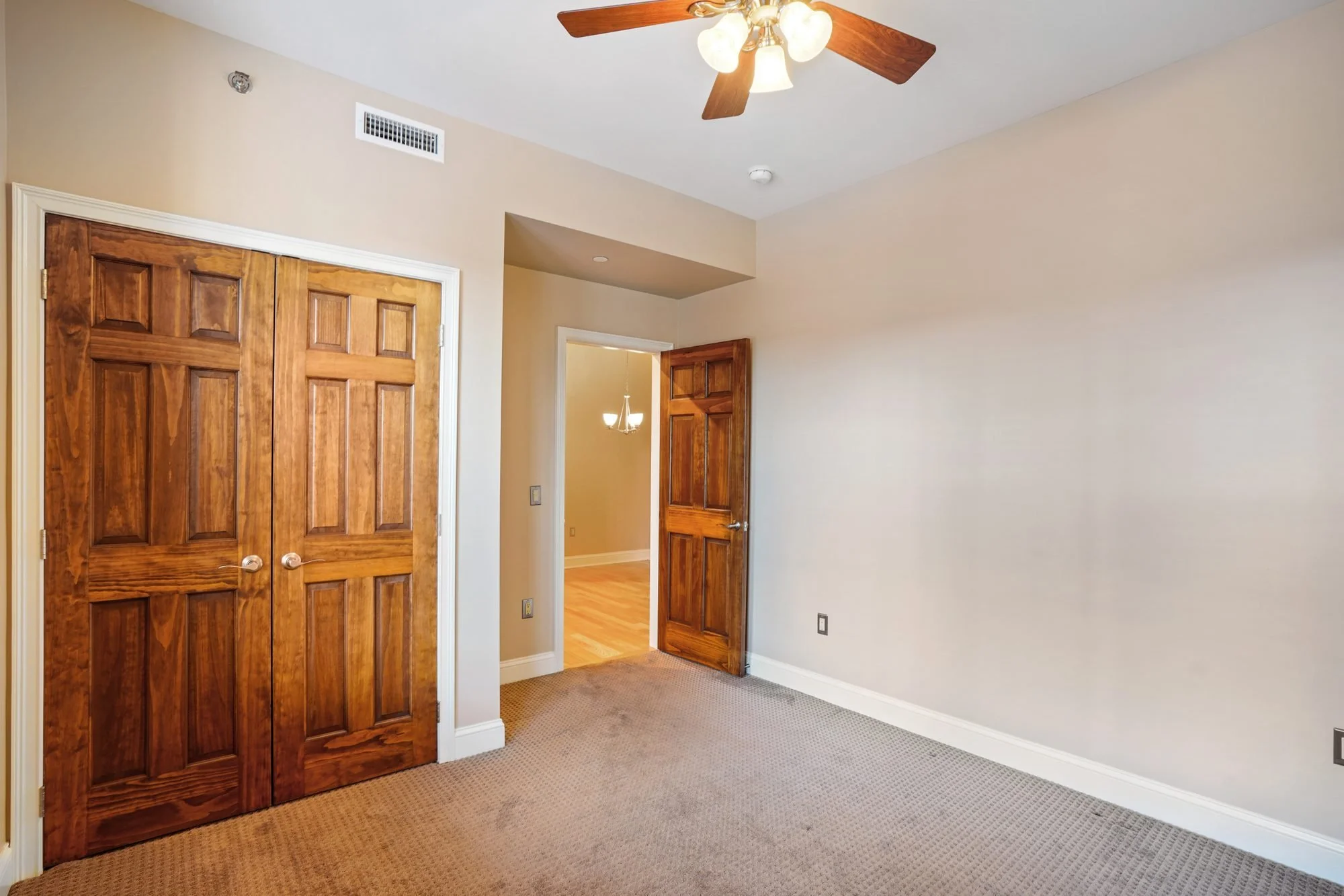 Empty room with beige walls, two wooden doors, carpeted flooring, ceiling fan with wooden blades and glass light fixtures, and a chandelier visible in an adjacent room