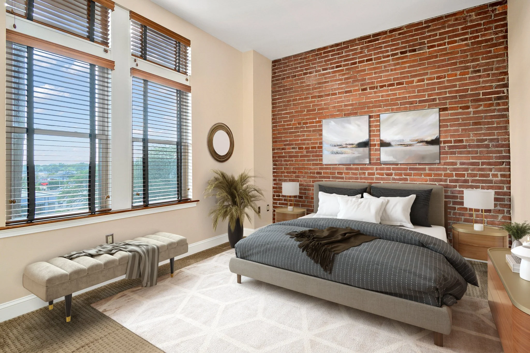 Modern bedroom with exposed red brick wall, large windows with blinds, a bed with gray and white bedding, two nightstands with lamps, a beige bench, and wall art.