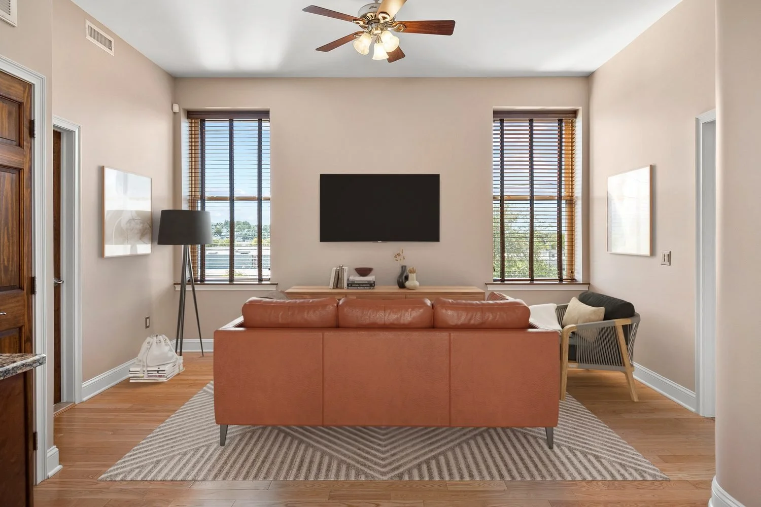 Living room with beige walls, hardwood floors, a leather sofa, a black floor lamp, a wooden sideboard, a wall-mounted TV, and two large windows with wooden blinds.