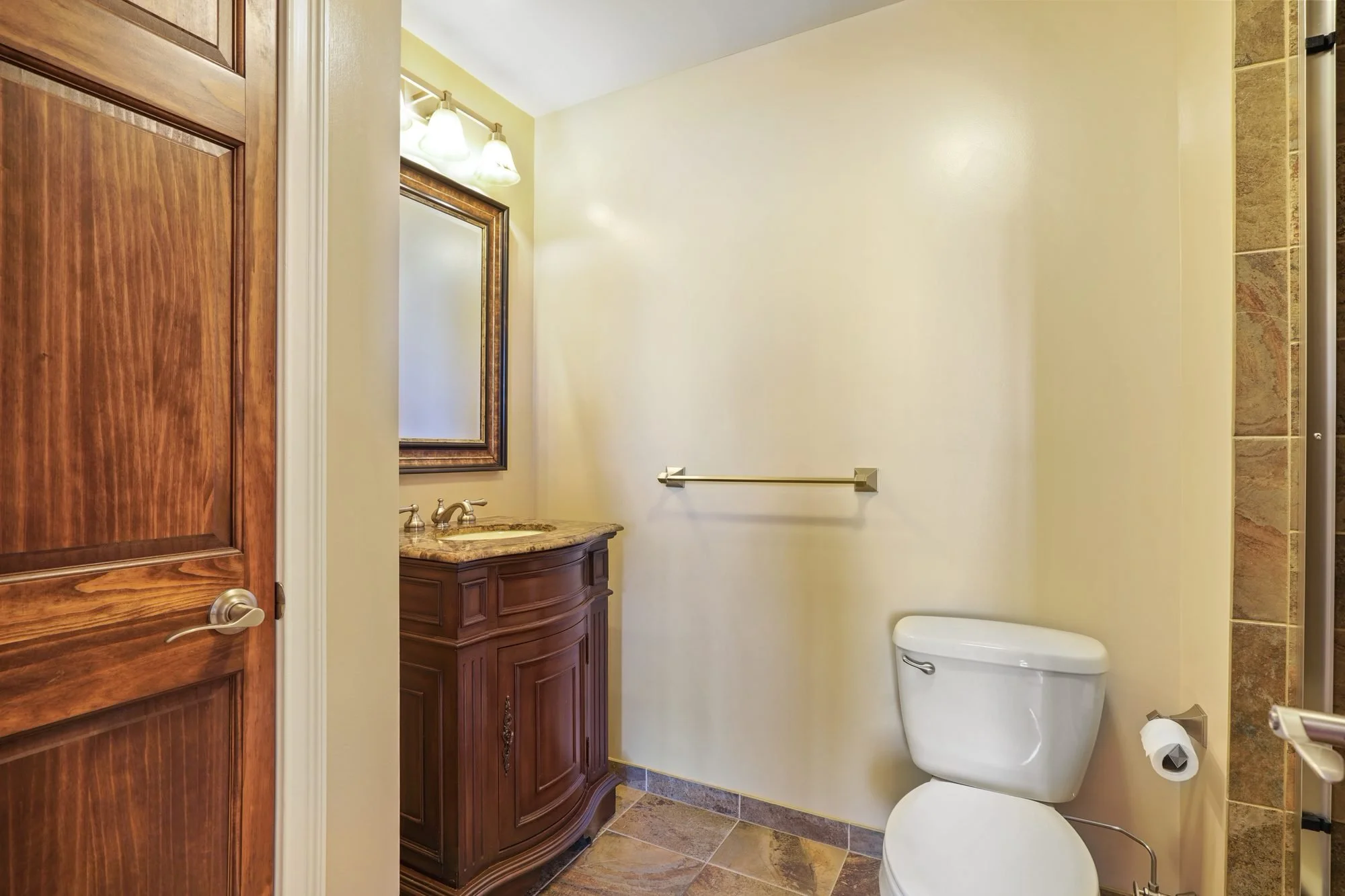 A small bathroom with a wooden vanity, mirror, and light fixture. There is a white toilet, a beige wall, and partial tiled shower area visible.