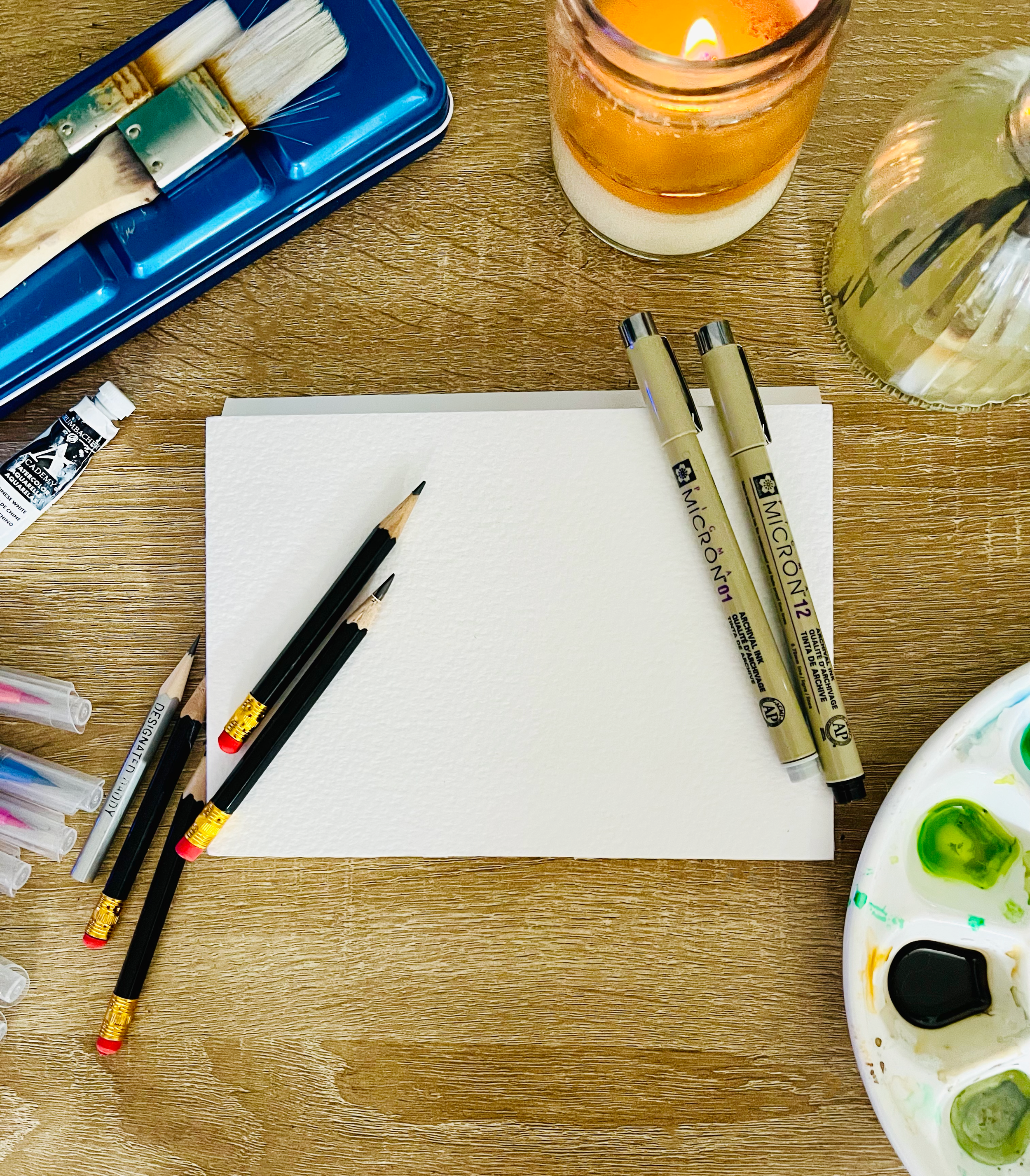 Art supplies on a wooden table including watercolor paints, paintbrushes, pens, and a lit candle.