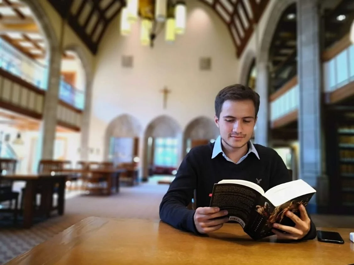 A young man reading a book at a wooden table inside a library or church.
