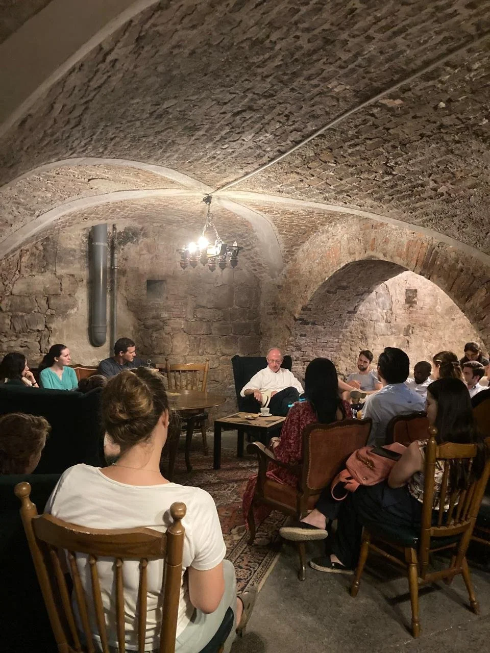 A group of people seated in a circular arrangement inside a stone basement with arched ceilings, listening to a speaker who is sitting at the center.