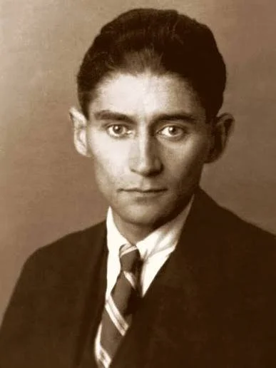 A black-and-white portrait of a young man with neatly combed hair, wearing a dark suit, white shirt, and tie, looking directly at the camera.