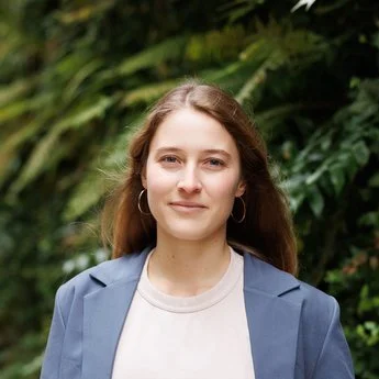 Young woman with long brown hair wearing a blue blazer and white shirt, standing outdoors with green foliage in the background.