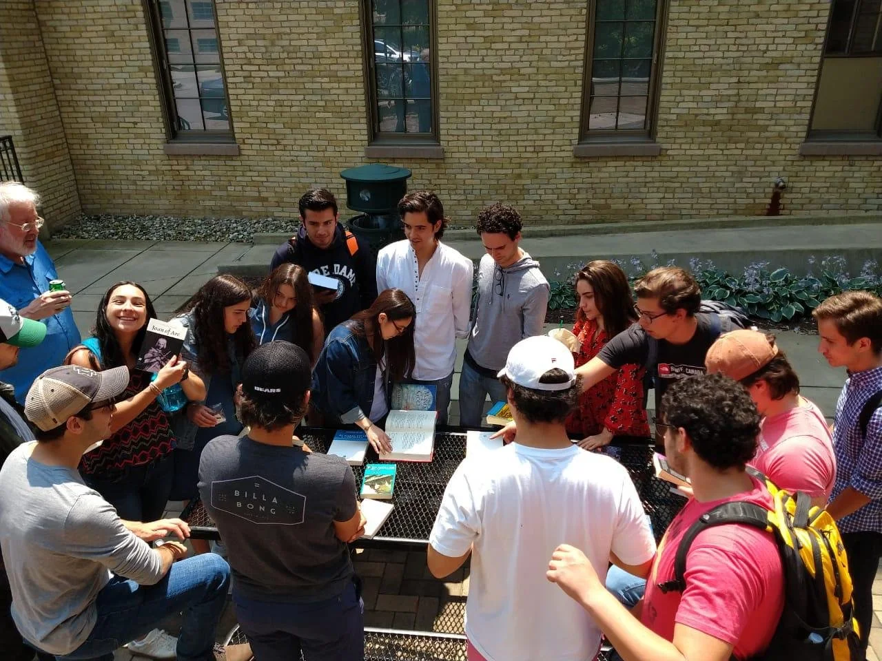 A group of young people gathered around a table outside, looking at books and magazines, with some pointing and engaging in conversation. An older man is standing on the left side, holding a drink, and several people are wearing backpacks and casual clothes.