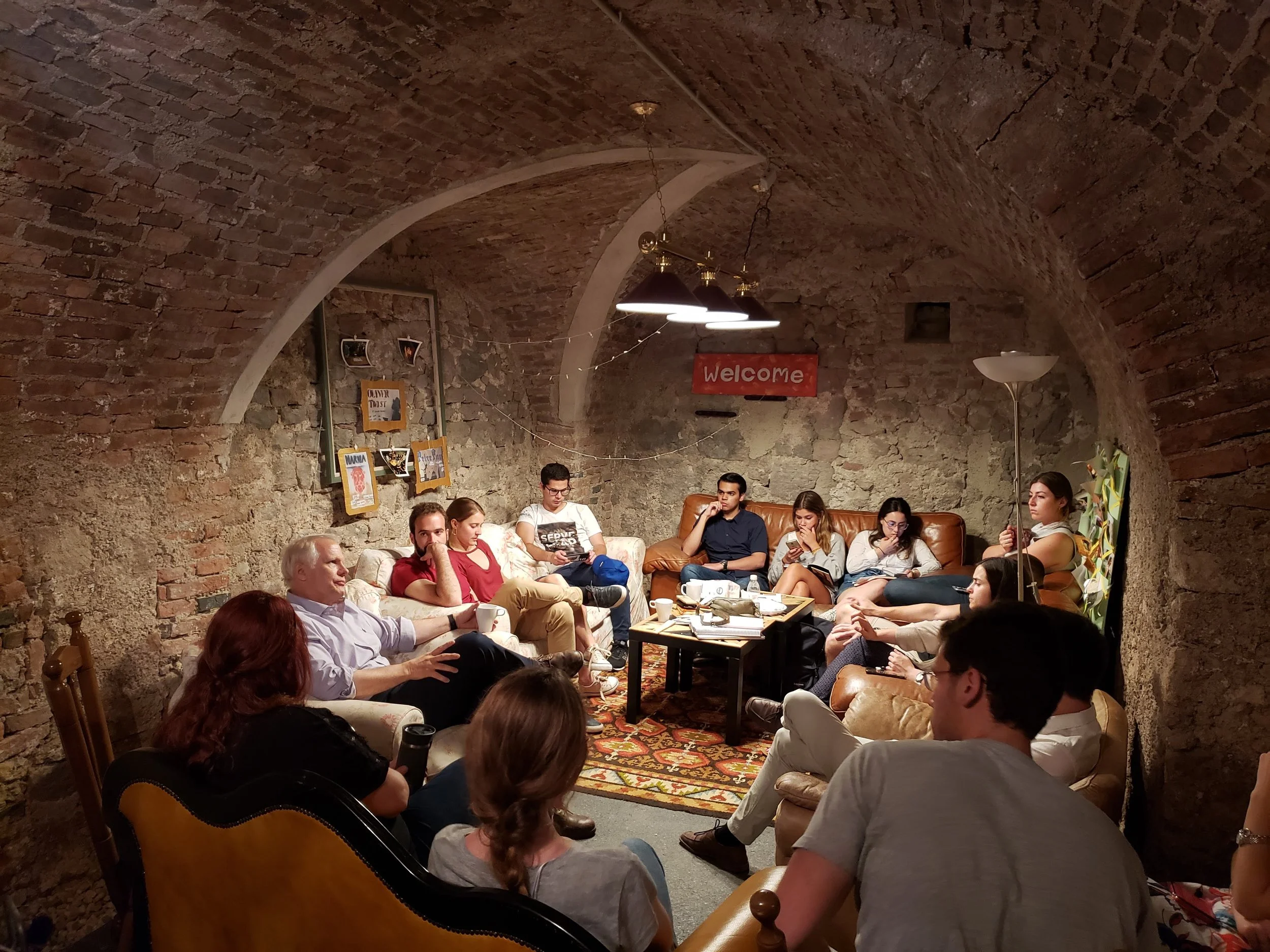 A group of people sitting and talking in a basement or cellar with exposed brick and stone walls, arched ceilings, and warm lighting. There is a