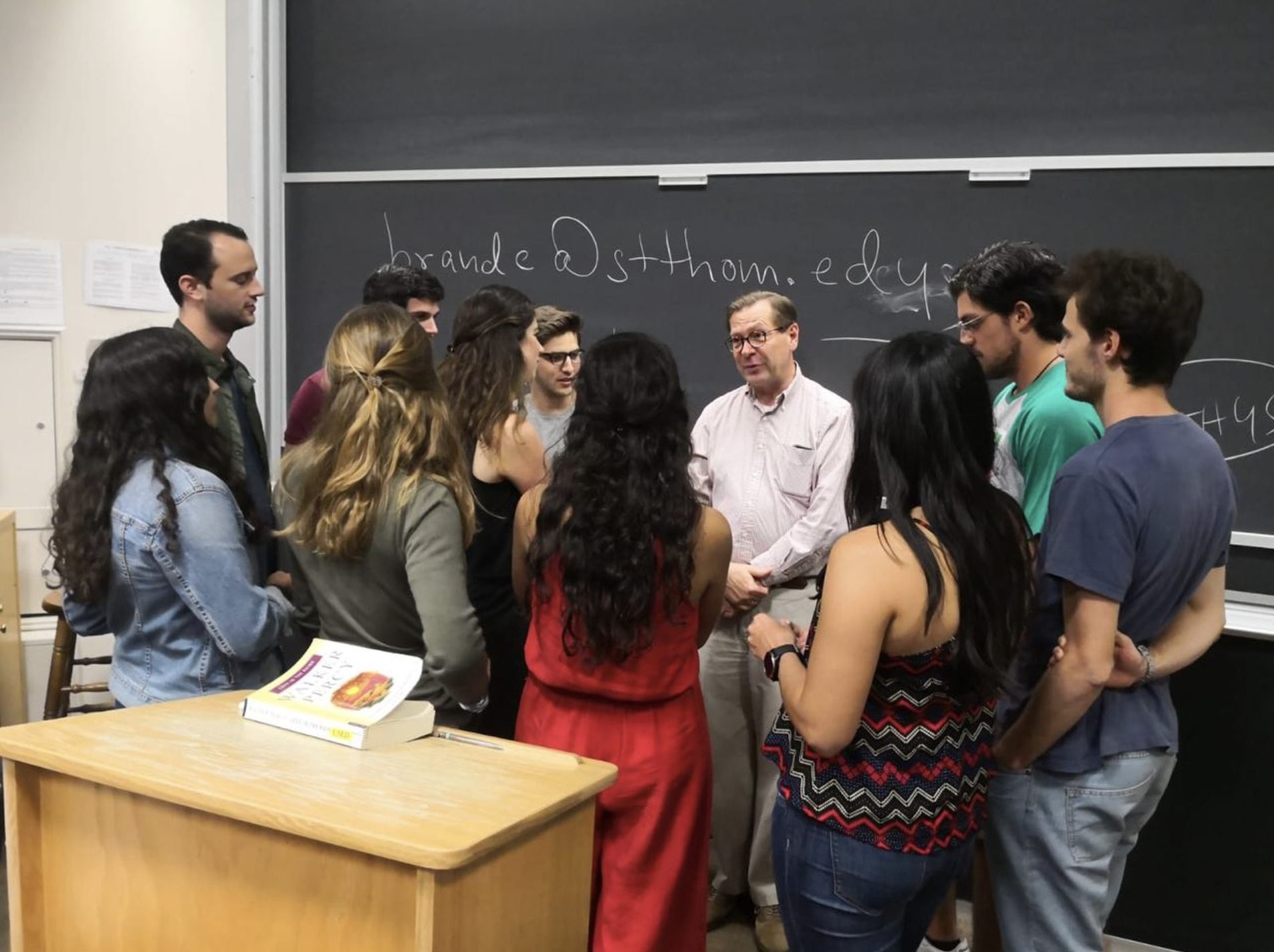 A group of students and a professor standing and talking in a classroom near a chalkboard with writing on it.