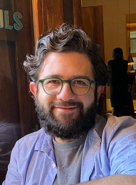 A smiling man with glasses, a beard, and wavy hair, sitting indoors with wooden walls and a person in the background.