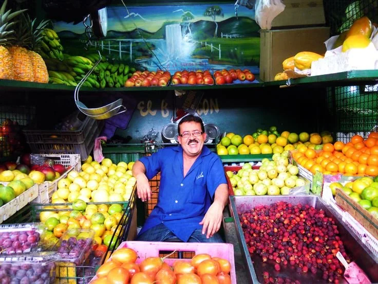 Hombre sonriendo en puesto de frutas, rodeado de manzanas, duraznos, uvas, y berries en un mercado