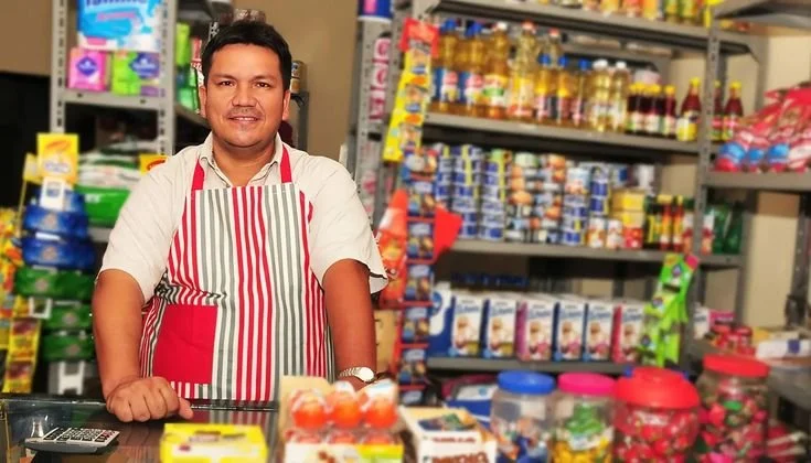 Hombre sonriendo en una tienda de abarrotes con un uniforme a rayas, rodeado de productos en los estantes.