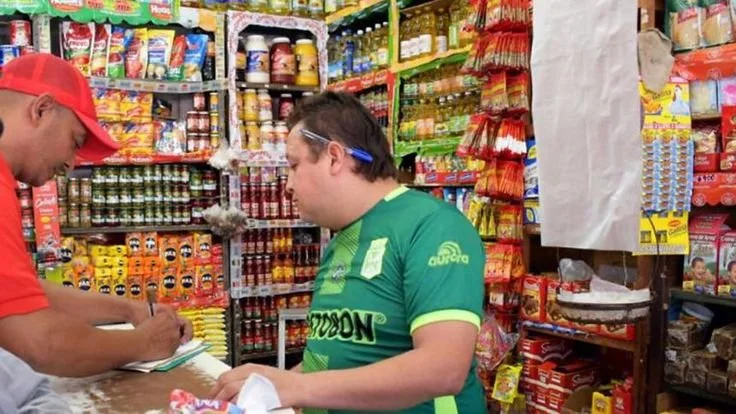 Dos hombres en una tienda de abarrotes, con estantes llenos de productos como condimentos, chicles y otros artículos de consumo diario.