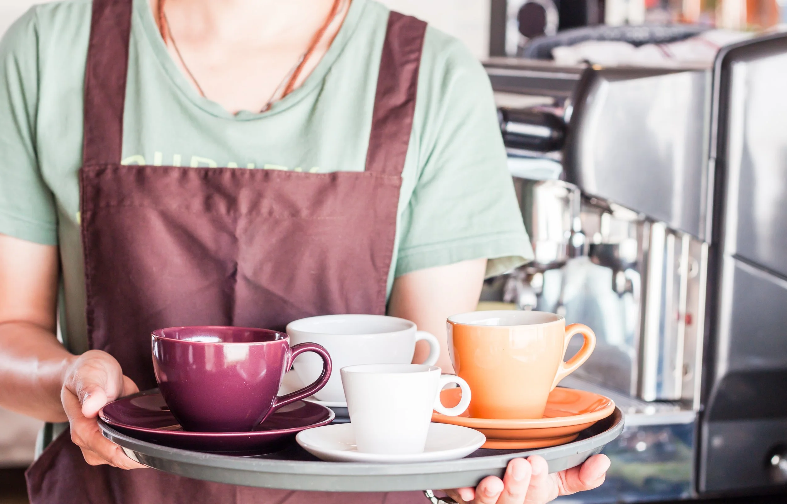 Persona sirviendo una bandeja con tazas de diferentes colores en un ambiente de cafetería.