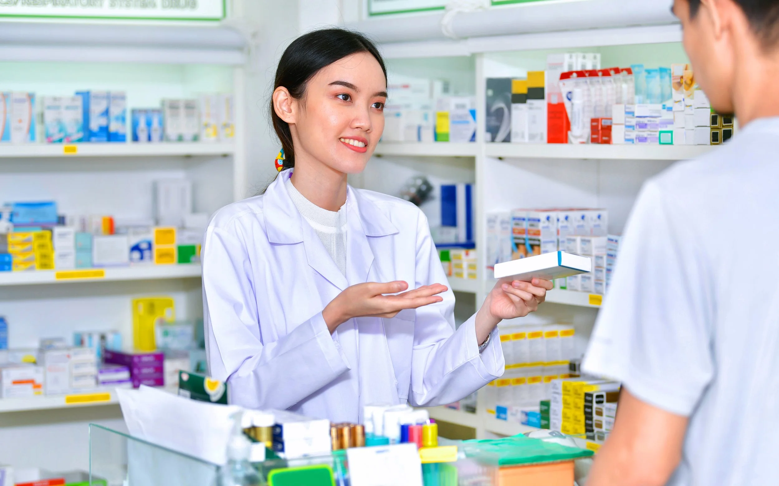 Una farmacéutica en bata blanca entregando una caja de medicamento a una clienta en una farmacia.