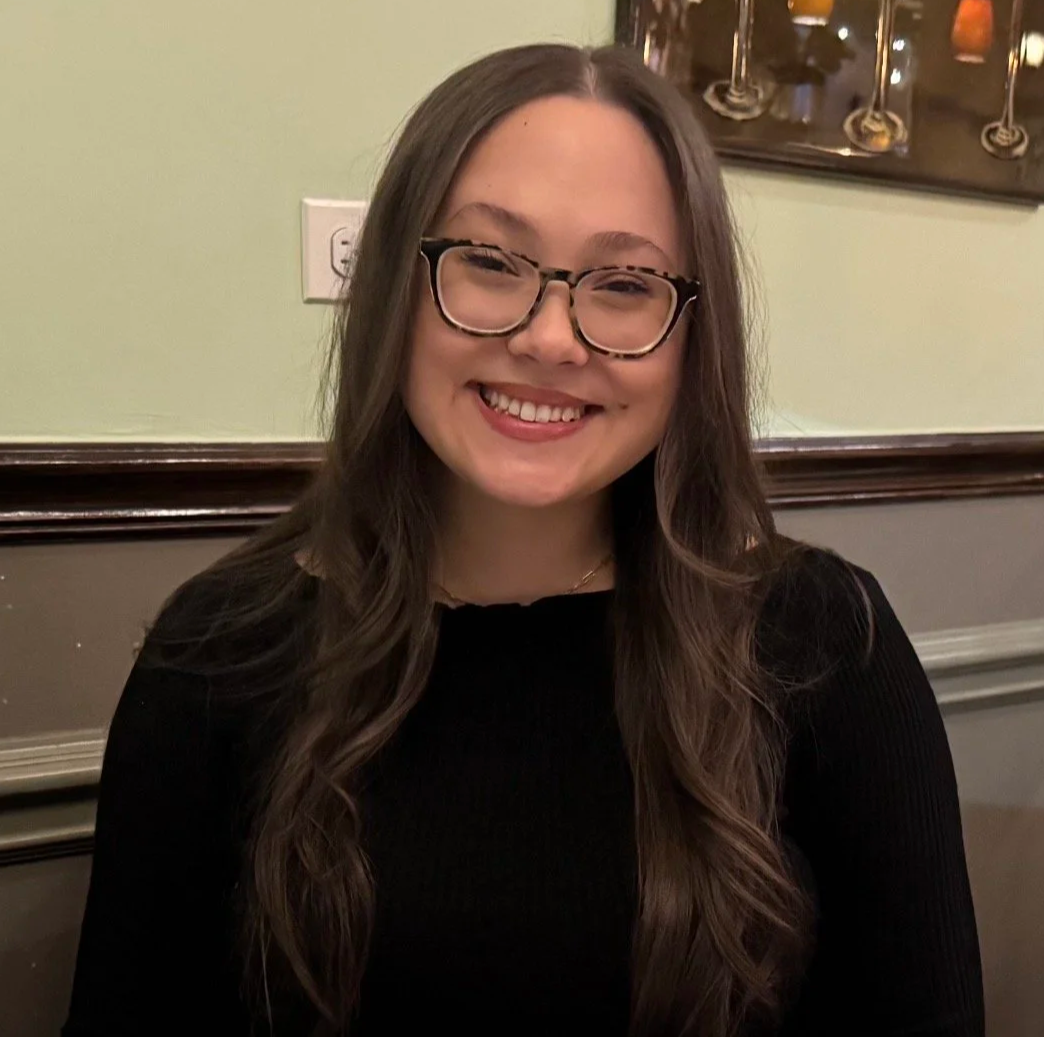 Ashlee Tarro, executive assistant to Dileesa. She is a White woman with long brown hair and glasses, smiling, wearing a black long sleeve shirt, sitting in a room with warm lighting.