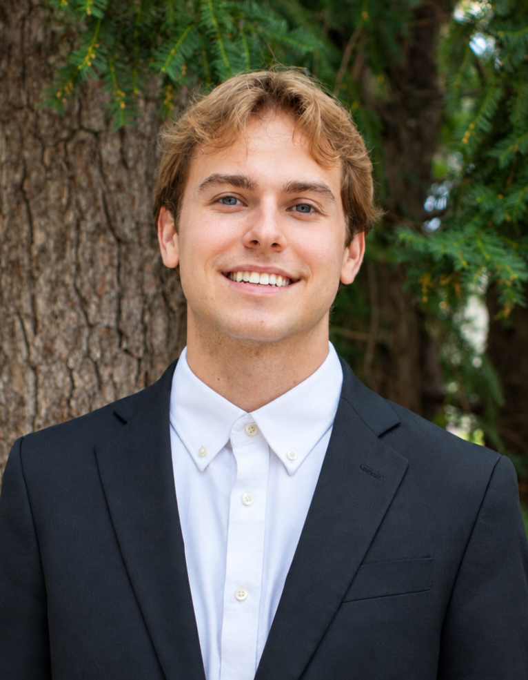 A young man in a formal black suit and white shirt standing outdoors in front of a tree with green foliage, smiling at the camera.