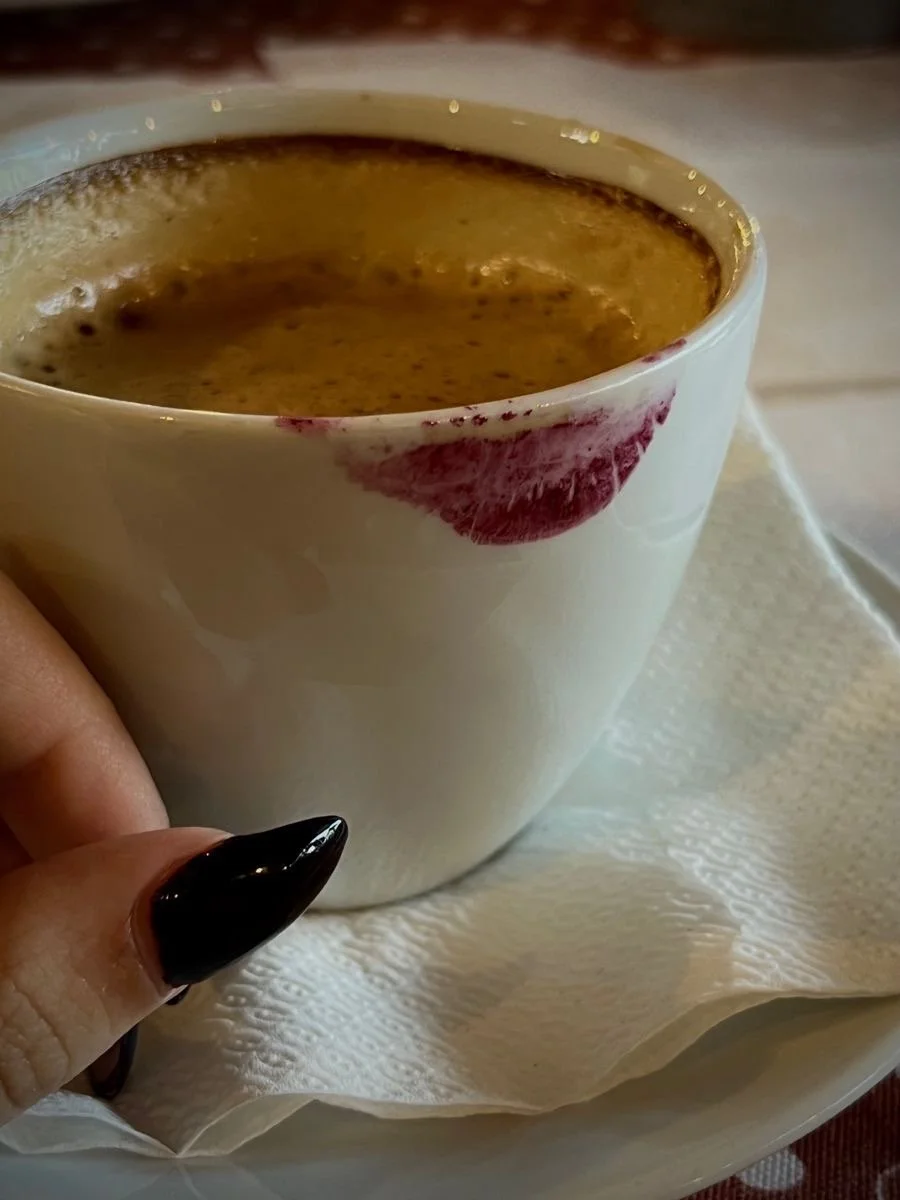 Close-up of a white coffee cup filled with black coffee, resting on a saucer with a paper napkin underneath. The cup has a lipstick mark on the rim. A hand with black-painted nails holds the cup.