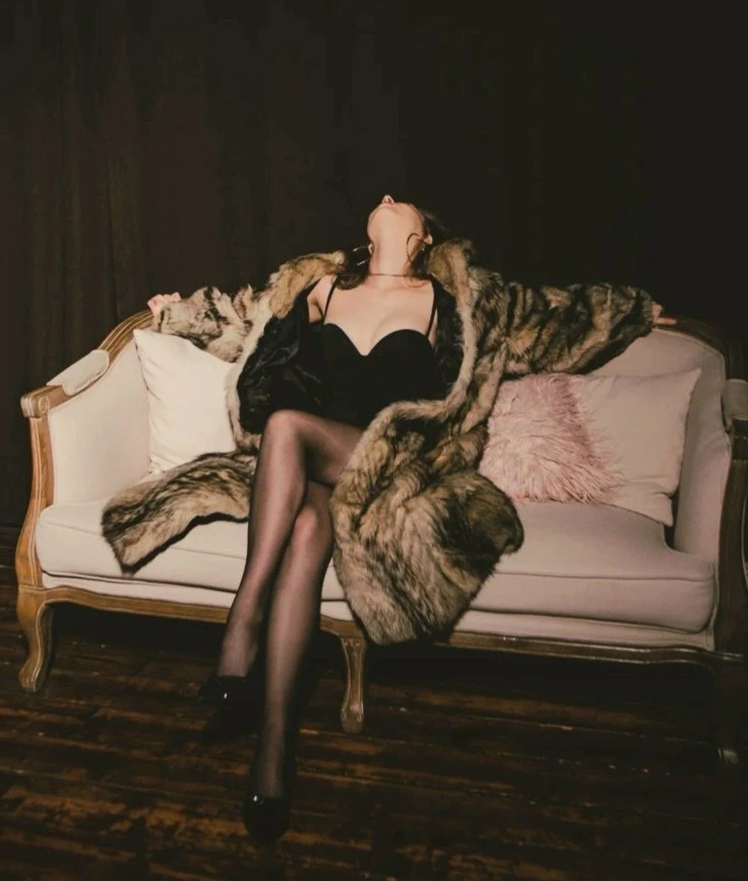A woman in black lingerie and sheer stockings sitting on a vintage sofa with a fur coat draped over her shoulders, her head tilted back and eyes closed.