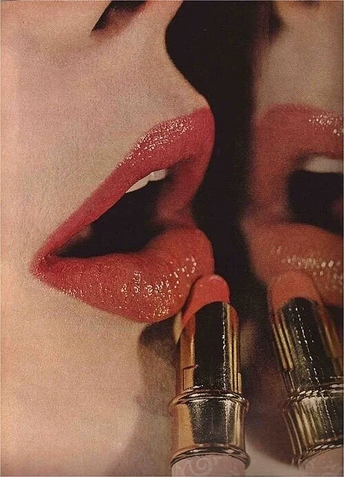 Close-up of a person's lips applying coral lipstick, reflected in a mirror with a lipstick tube nearby.