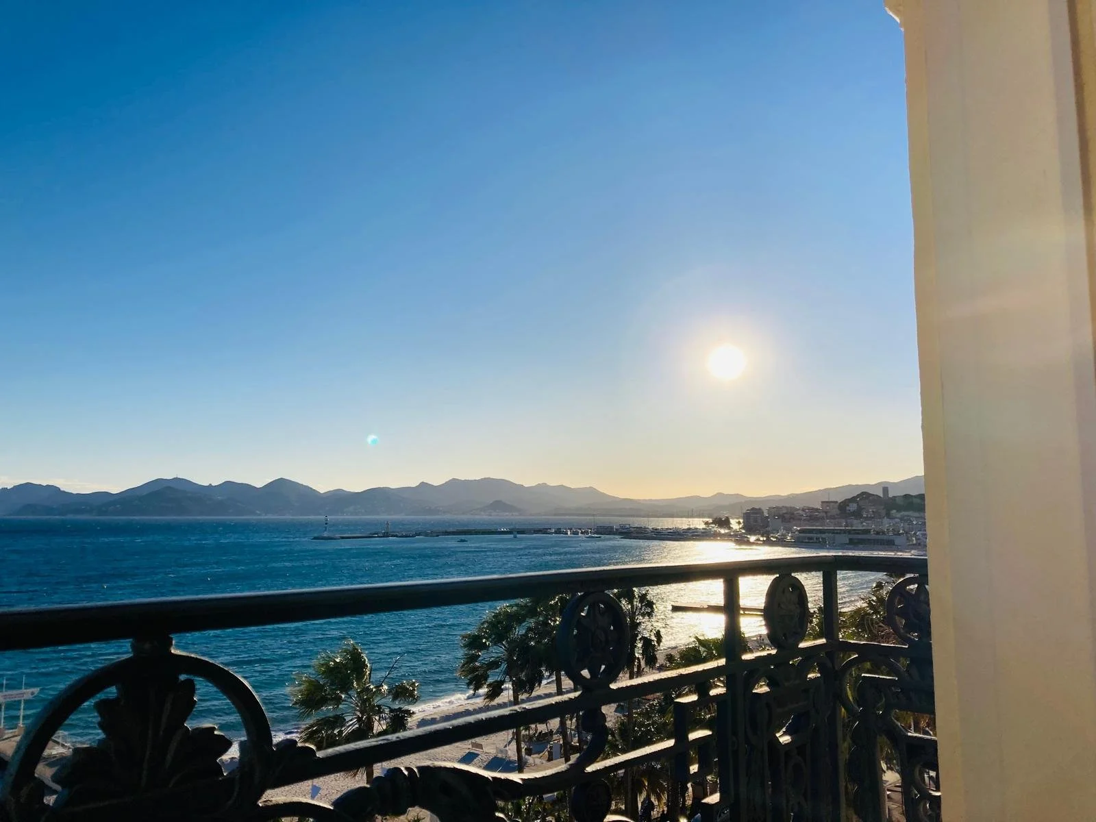 View of the ocean with mountains in the distance, a clear evening sky, and the sun setting, seen from a balcony with a decorative iron railing.