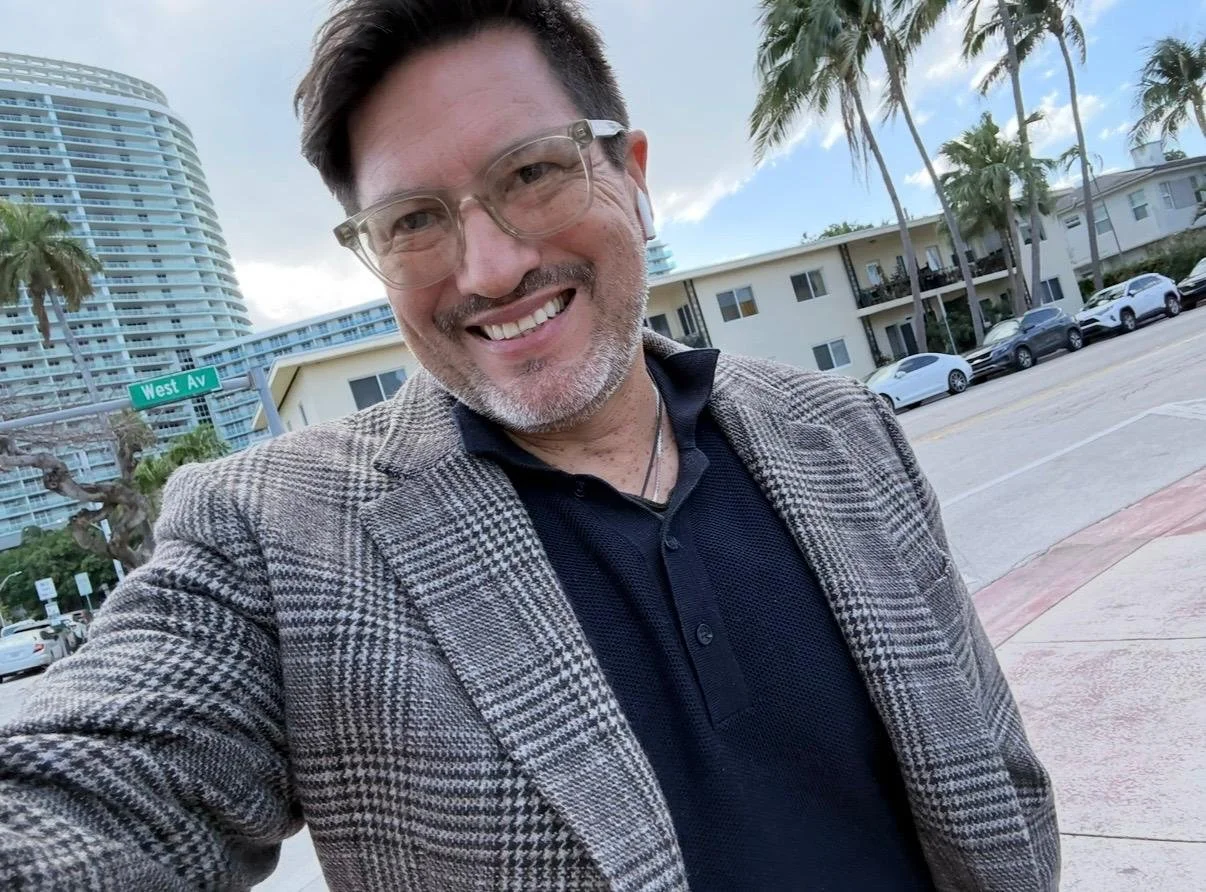 A smiling man taking a selfie outdoors near the street with buildings, palm trees, parked cars, and a street sign that reads 'West Av' in the background.