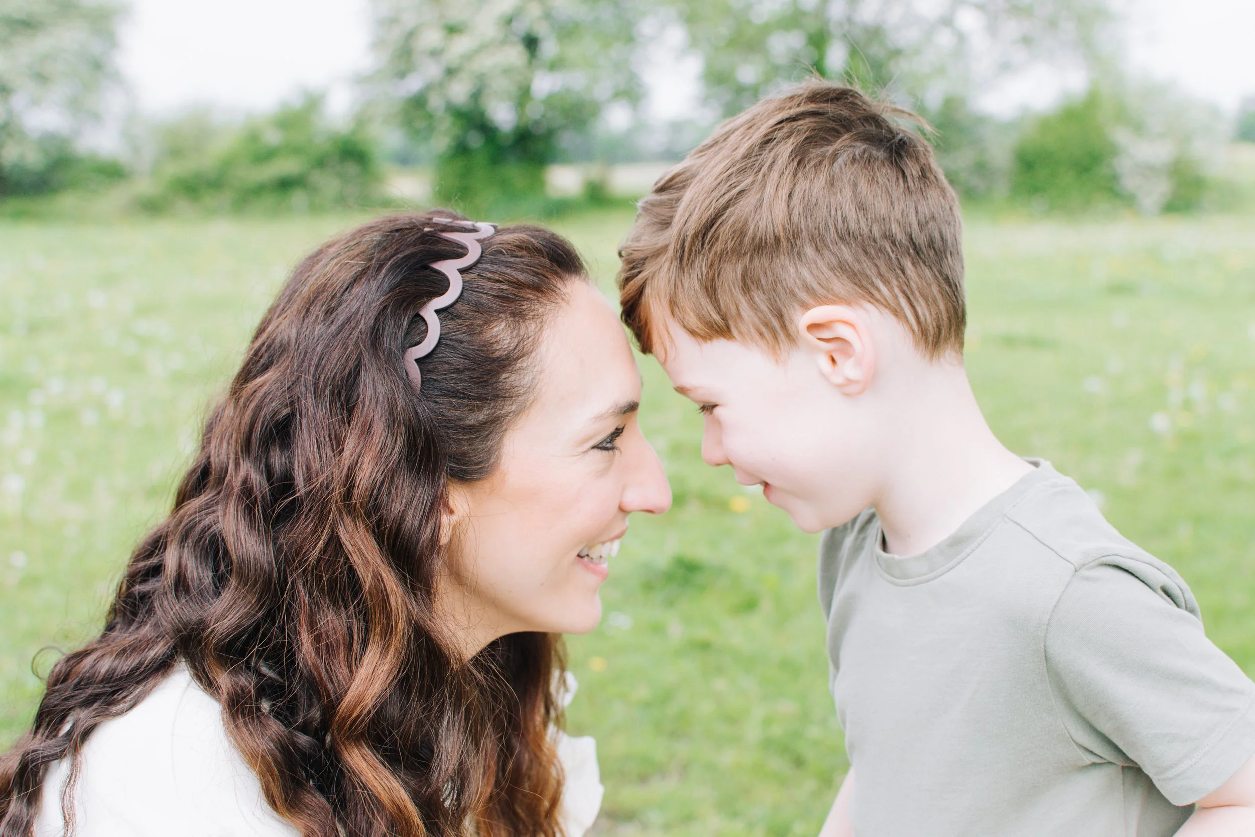 A woman and a young boy are smiling and touching foreheads in an outdoor park with green grass and trees in the background.