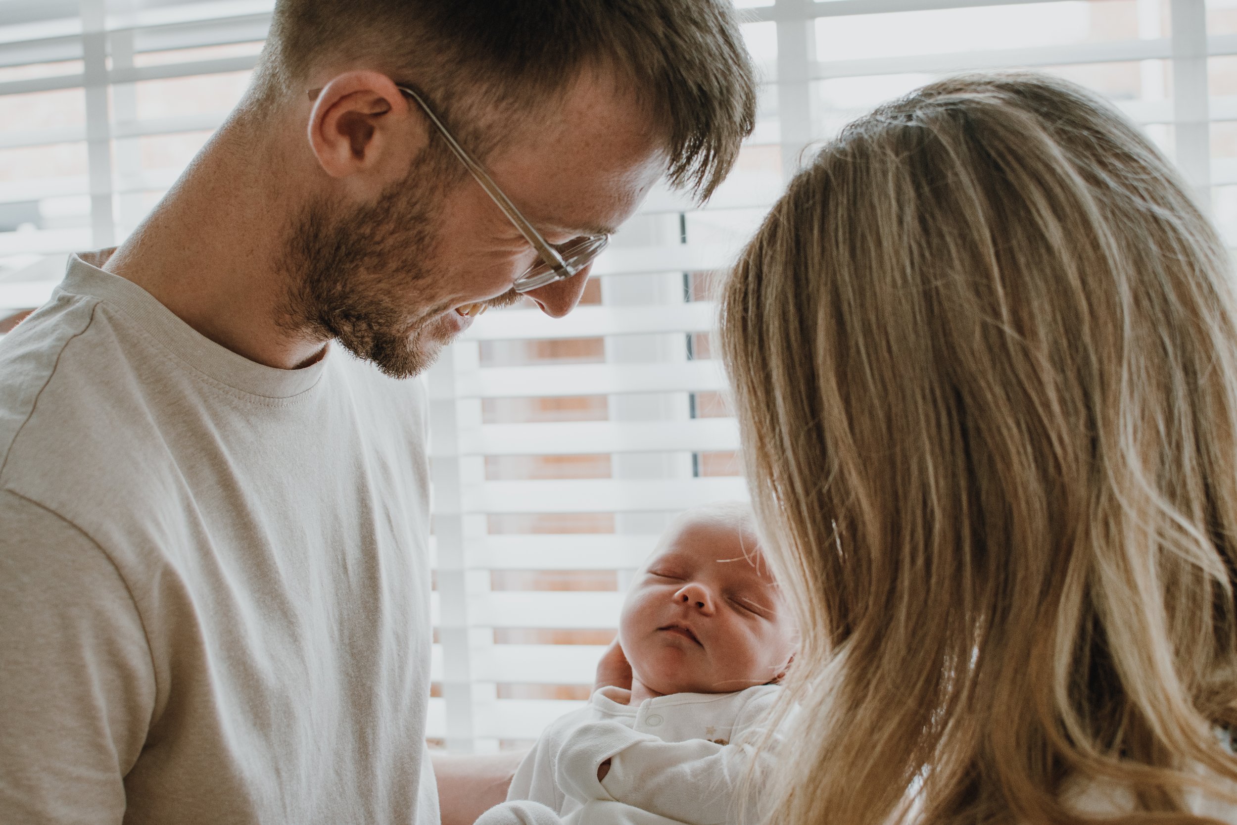 A man with glasses and a beard holding a newborn baby while a woman with long hair cradles the baby's head near a window with blinds.