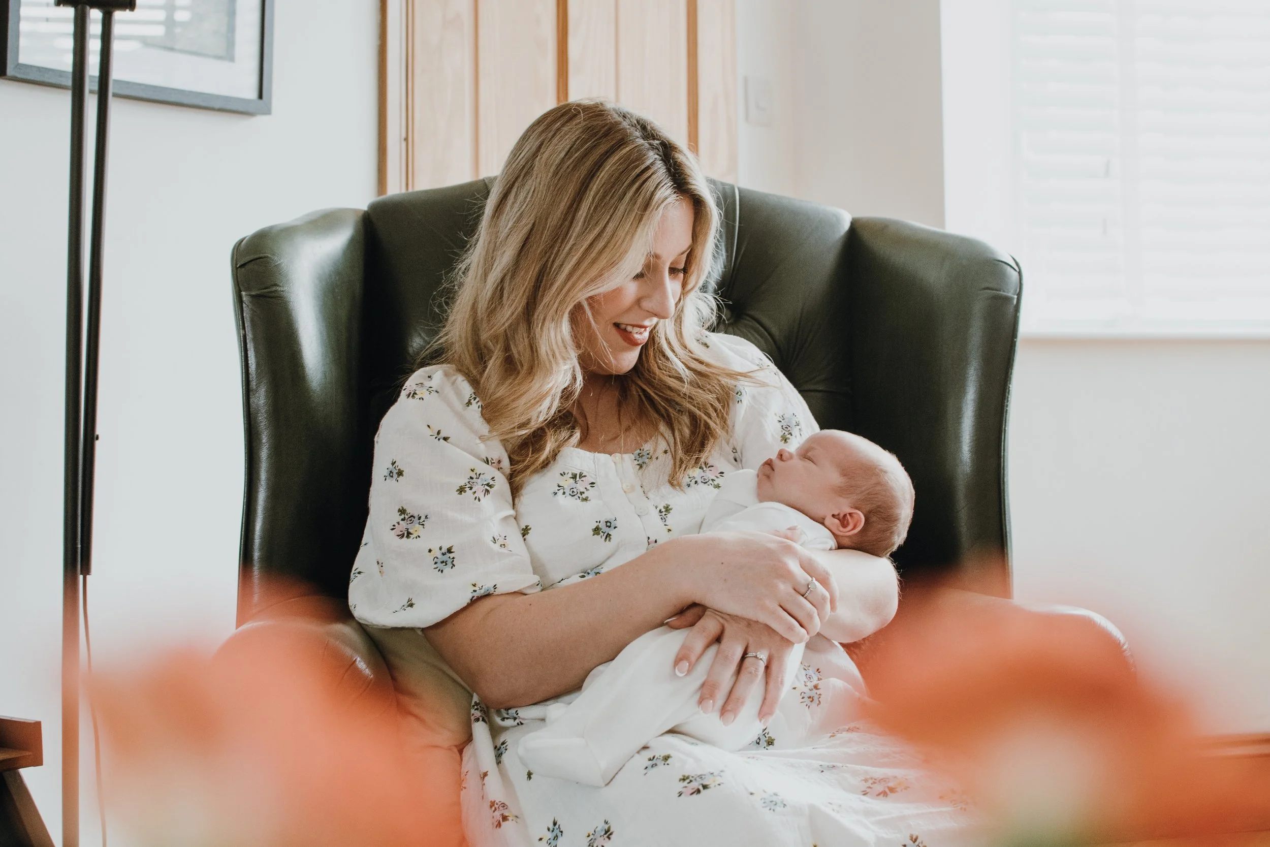 A woman with blonde hair smiling and holding a baby in her arms while sitting in a green armchair indoors.