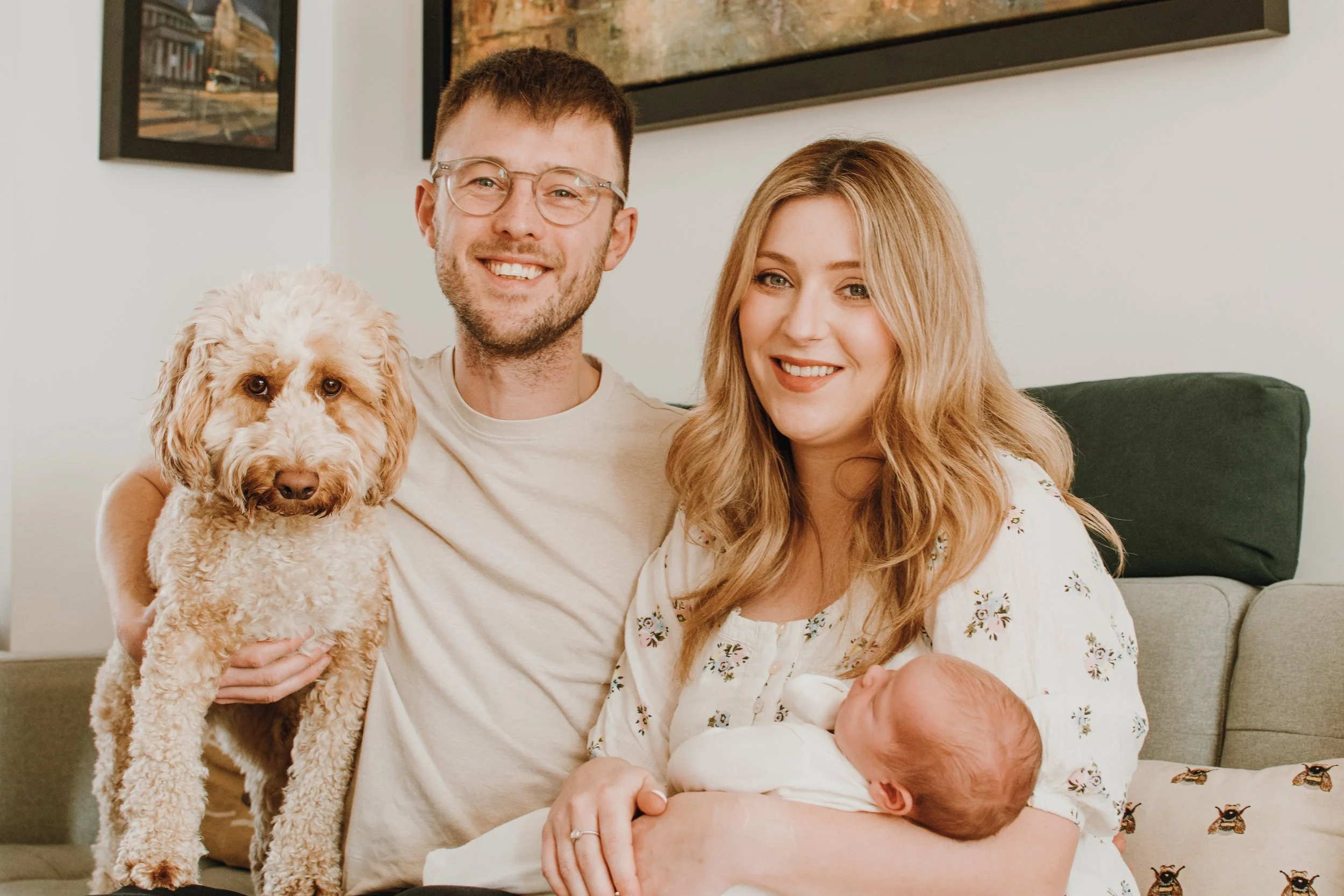 Family of four with a golden retriever dog. The man has short brown hair, glasses, and a beard, smiling and holding the dog. The woman has long wavy blonde hair, smiling, and a newborn baby wrapped in a blanket. They are sitting in a living room with