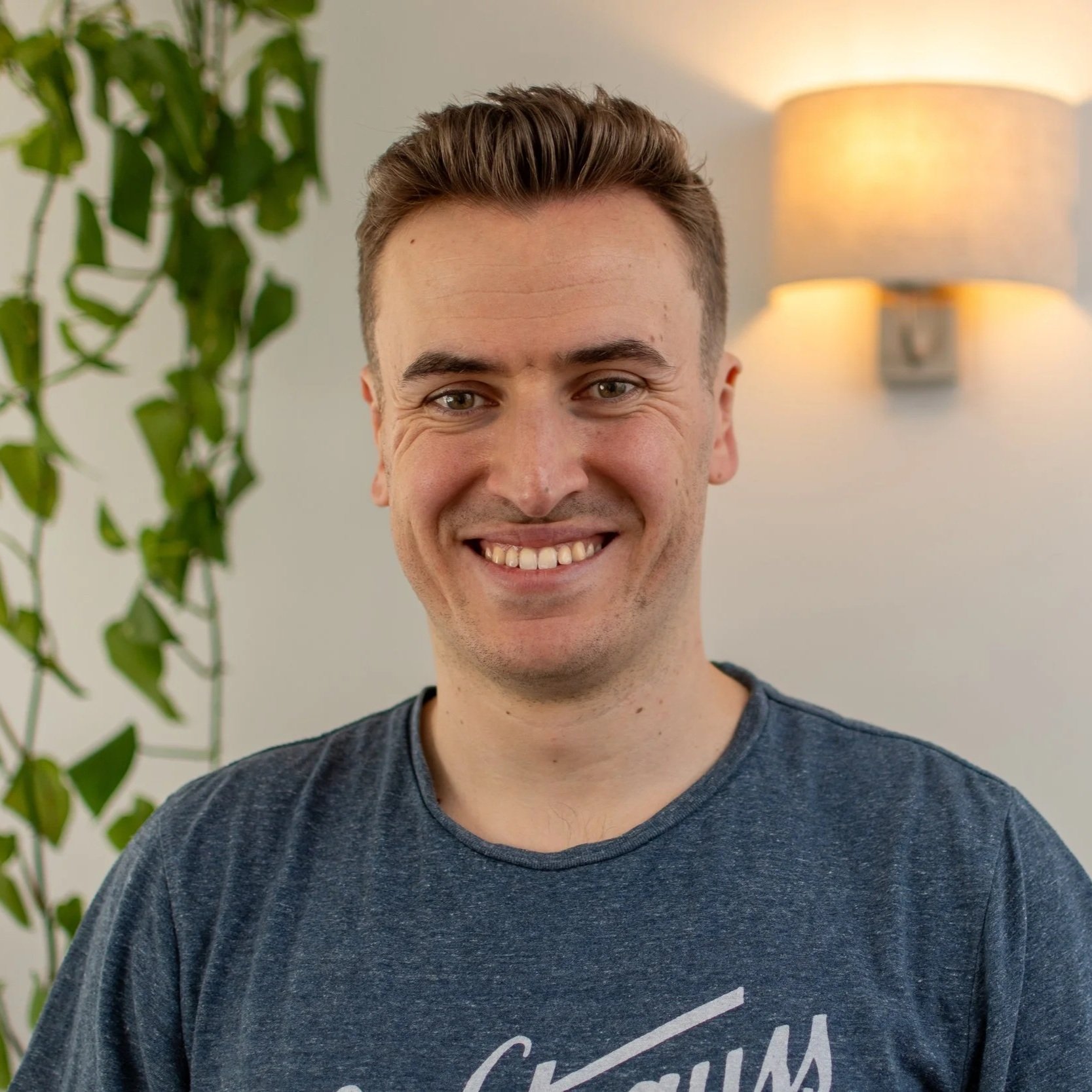 Young man smiling indoors with a plant and wall lamp in the background.