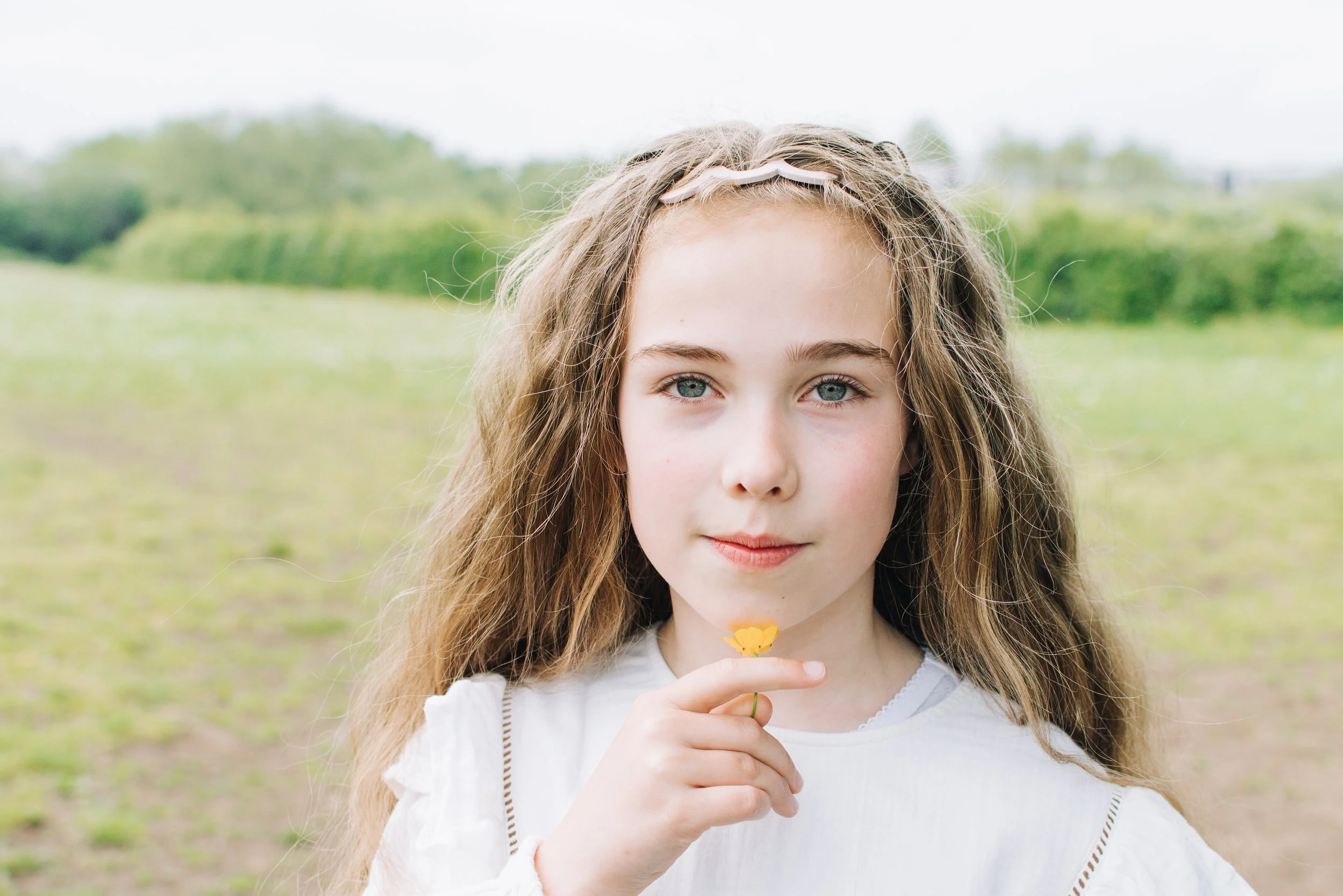 A young girl with long wavy brown hair and bright blue eyes holding a yellow flower, standing outdoors in a grassy field with green trees in the background.
