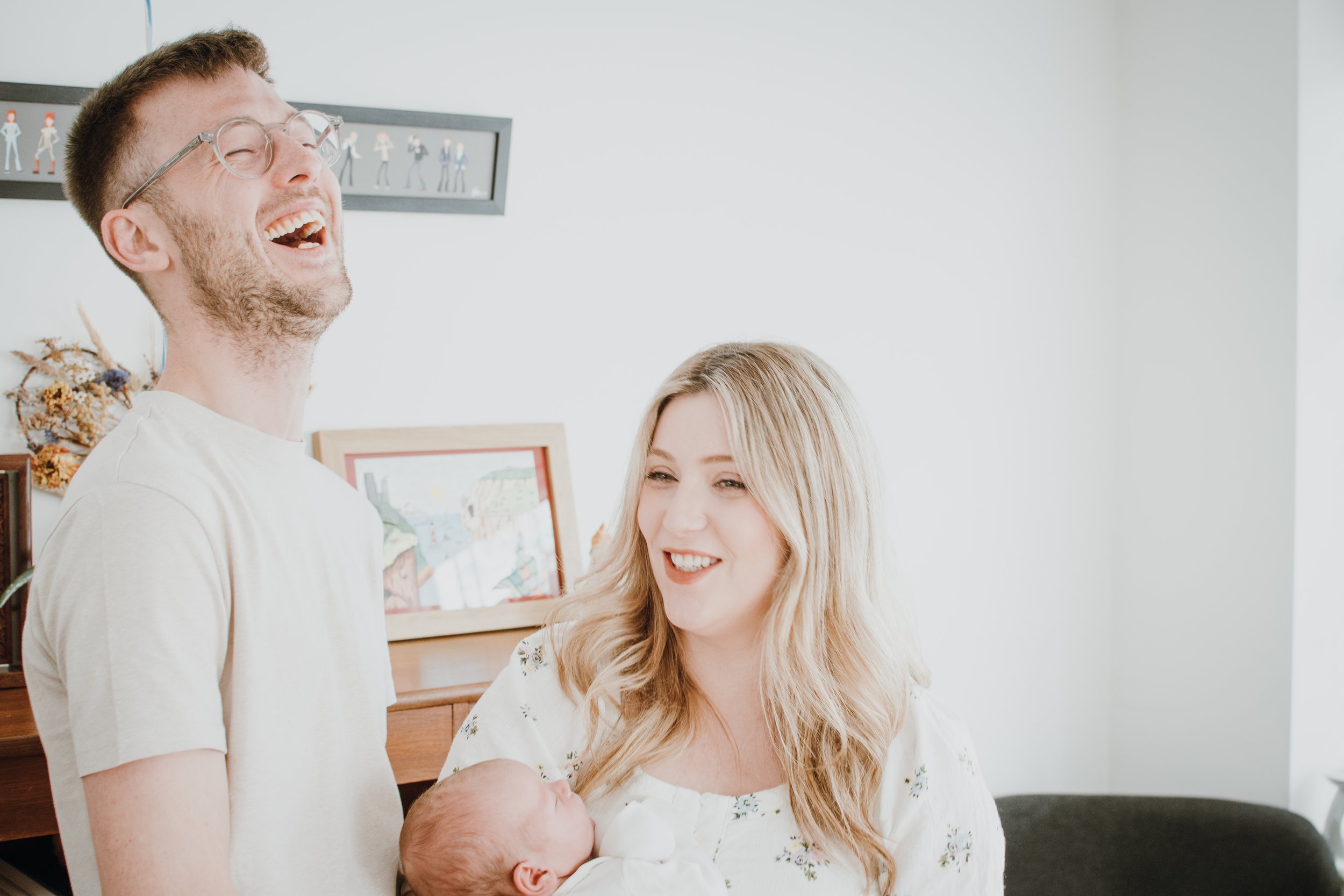 A happy man wearing glasses laughs while holding a newborn baby, with a woman smiling beside him in a bright indoor setting.