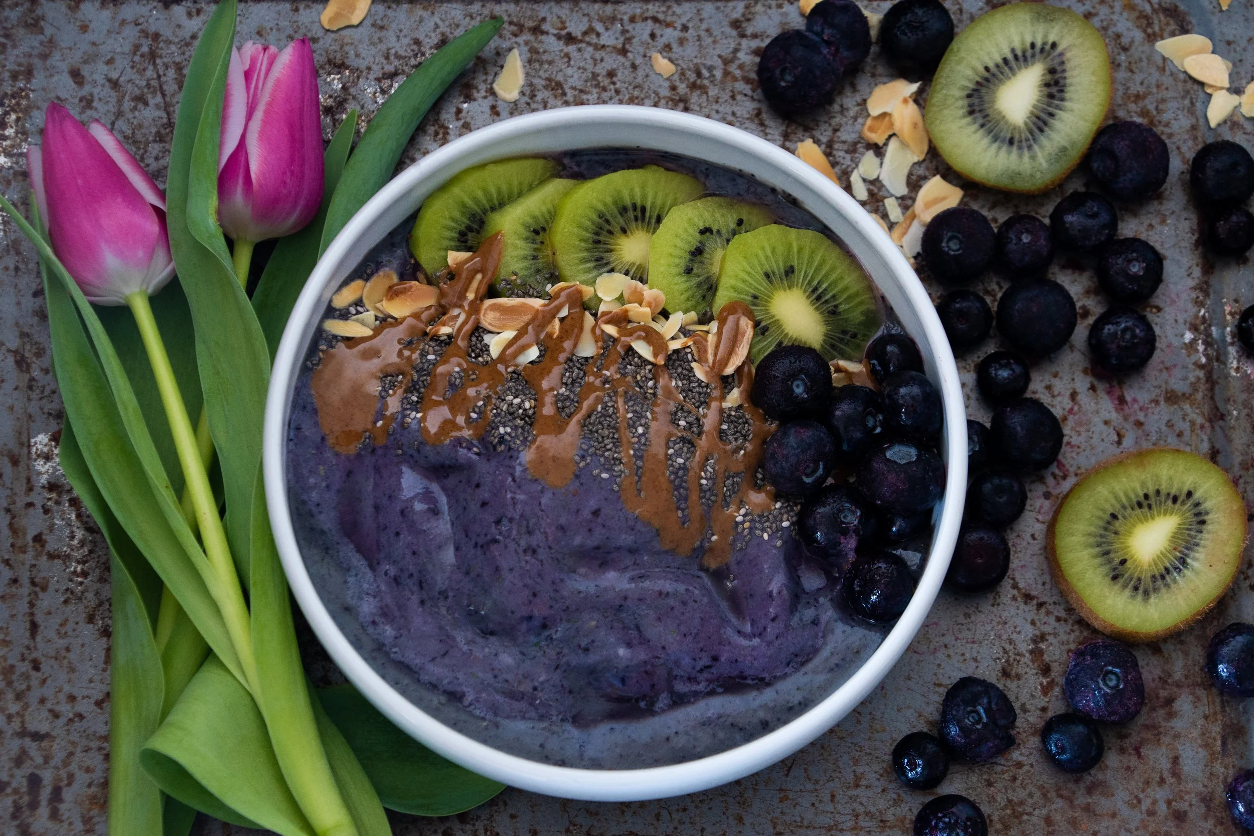 A bowl of purple smoothie topped with sliced kiwi, chopped almonds, and chia seeds, surrounded by blueberries and pink tulips on a rustic surface.