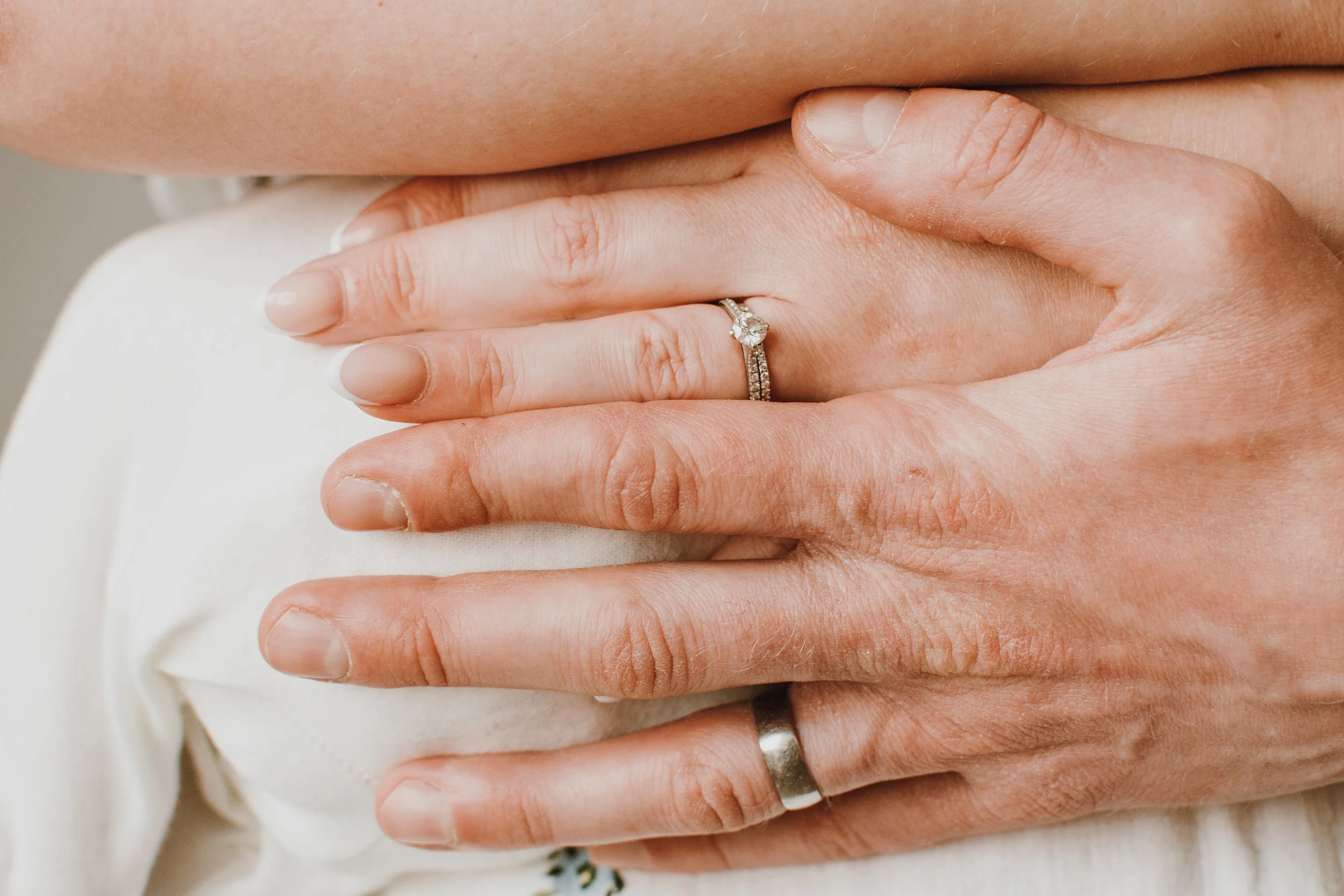 Close-up of hands, with two rings on the ring fingers, one featuring a large centerpiece gemstone and the other a band with small gemstones, suggesting a wedding or engagement ring. The hands are resting on a cream-colored fabric.