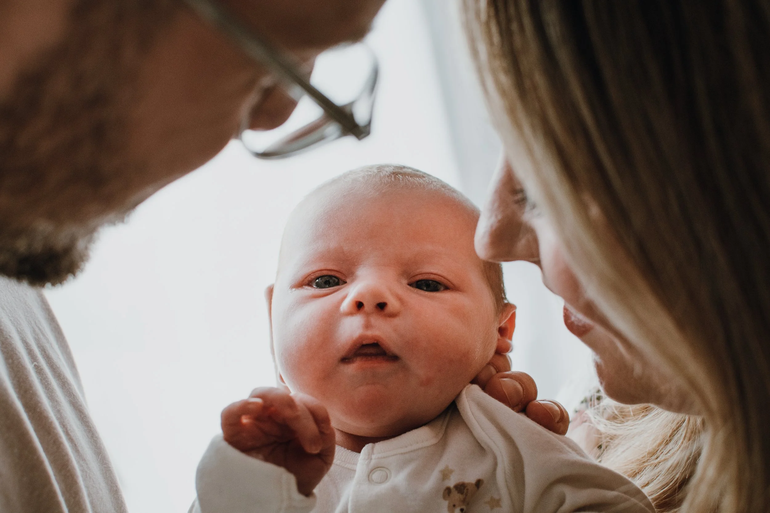 Close-up of a man and woman holding a baby, viewed from below. The baby's face is in focus, looking directly at the camera with blue eyes. The man and woman are leaning in close, with their faces near the baby's head.