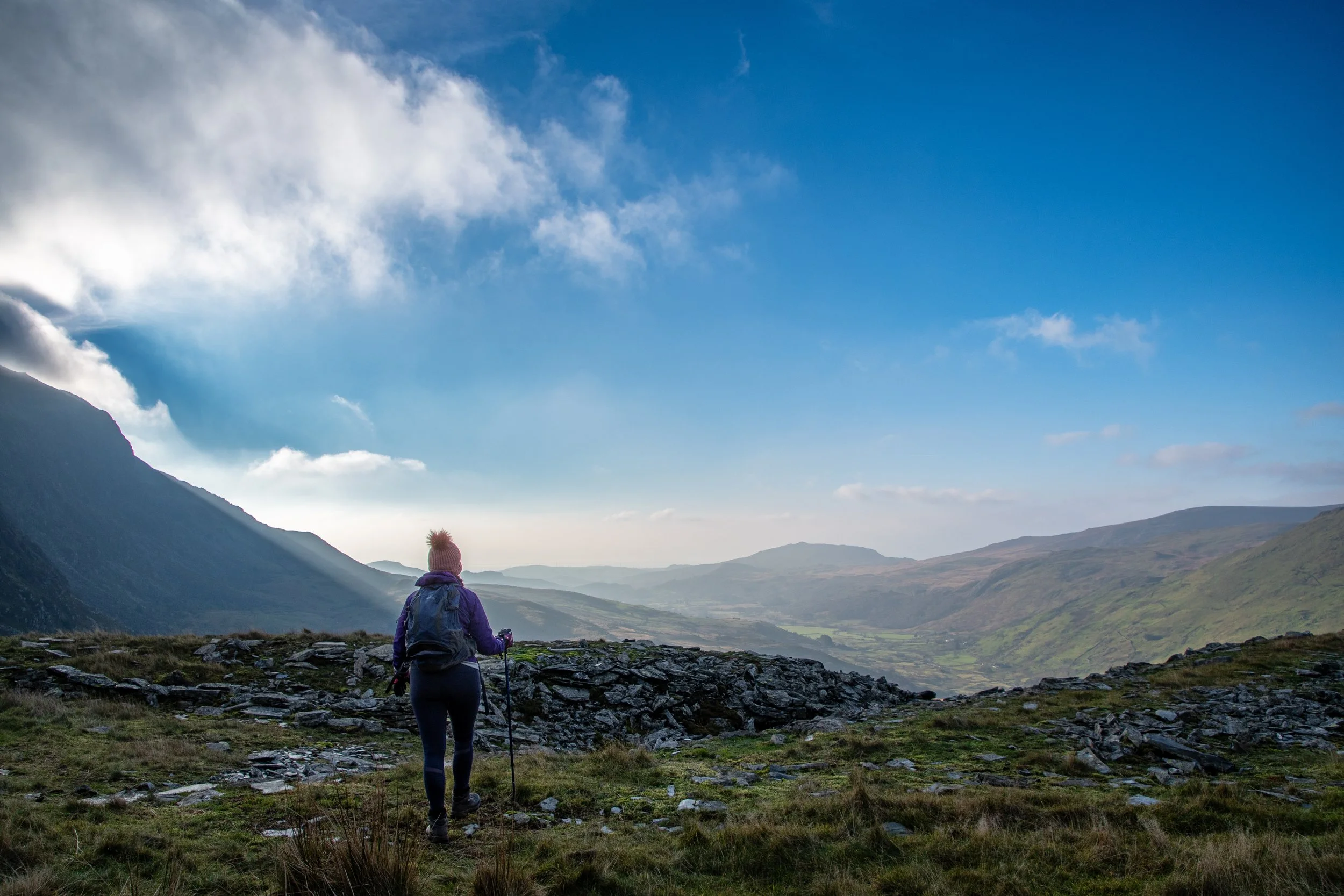 A person hiking in a mountainous landscape with rolling green hills, rocky terrain, and a partly cloudy sky.
