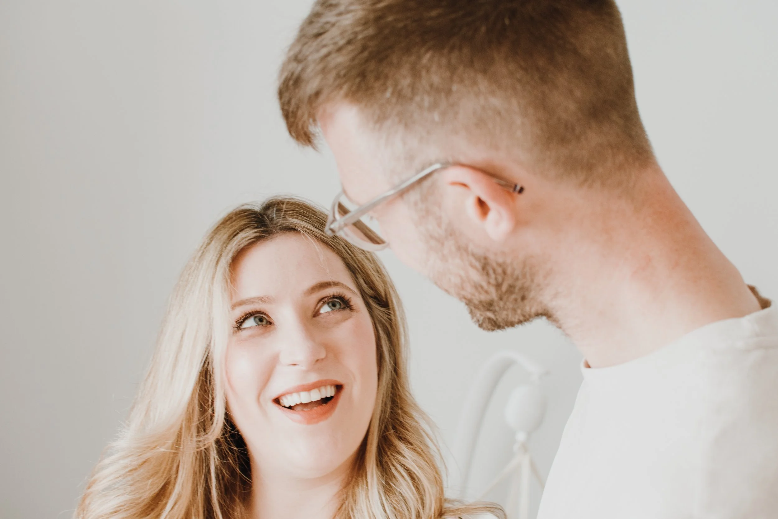 A man and woman smiling at each other, with the woman looking up at the man. The woman has long blonde hair and the man is wearing glasses and has short brown hair. They are both smiling and appear happy.