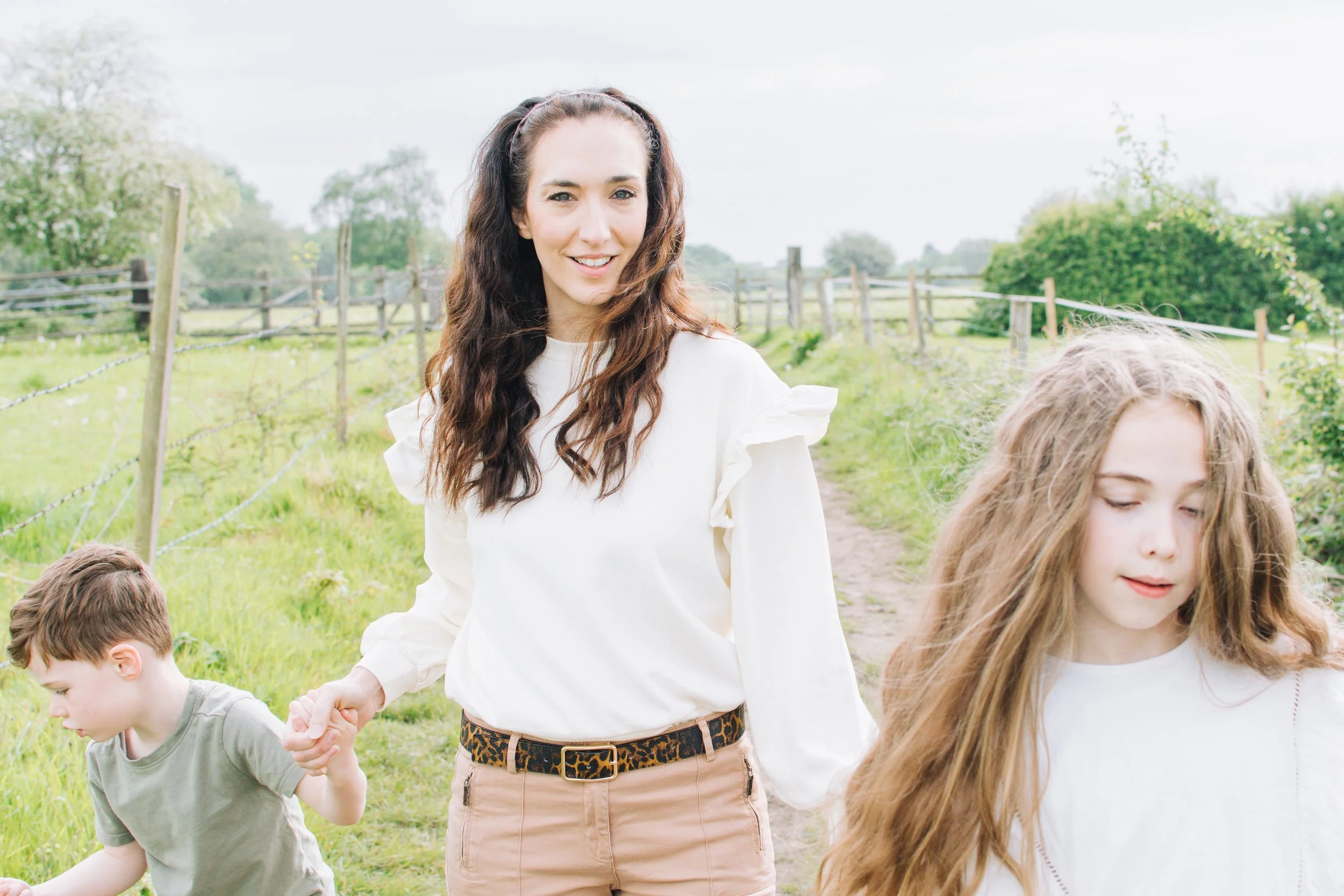 A woman with long wavy brown hair, wearing a white blouse and beige pants with a leopard print belt, holding hands and walking with children outdoors in a green field with trees and fencing in the background.