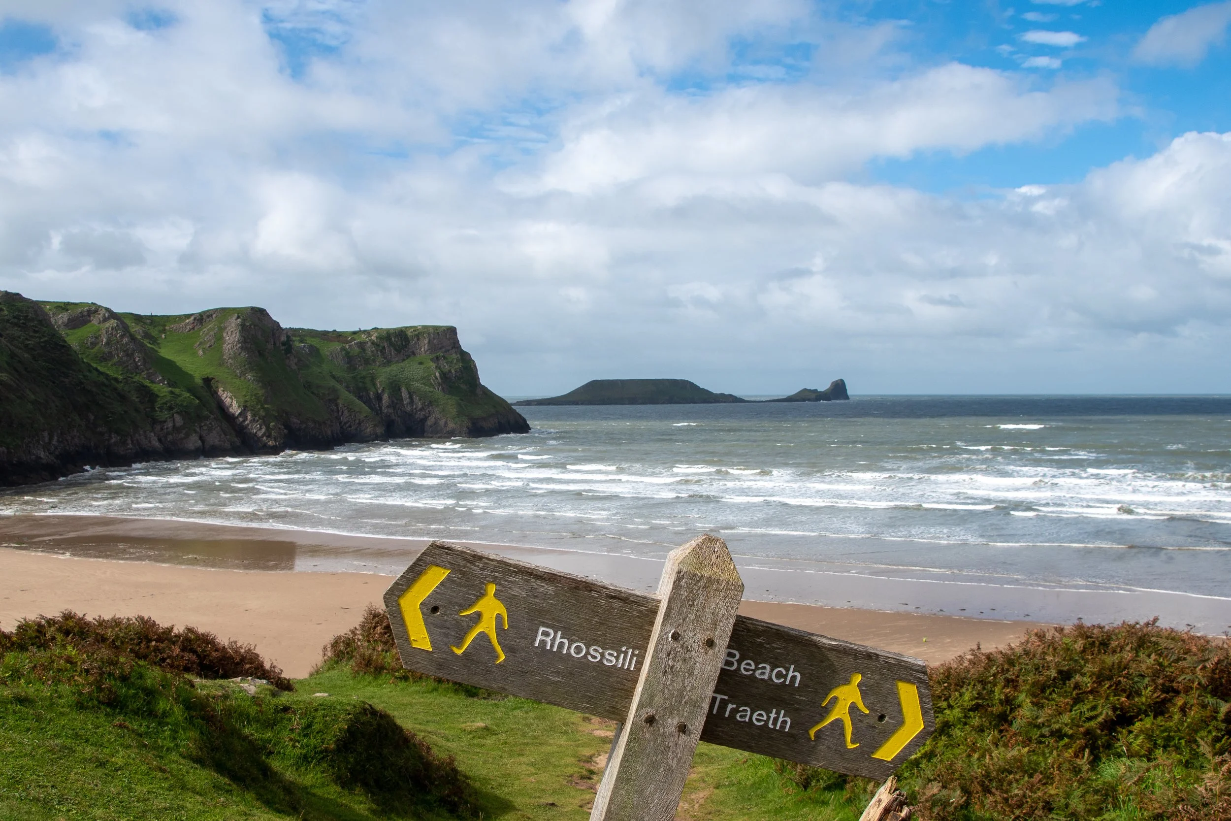 Photo of a beach with waves hitting the shore, green cliffs in the background, and a cloudy sky overhead. There is a sign in the foreground that reads 'Rhossili Beach Trail' with walking figures and directional arrows.