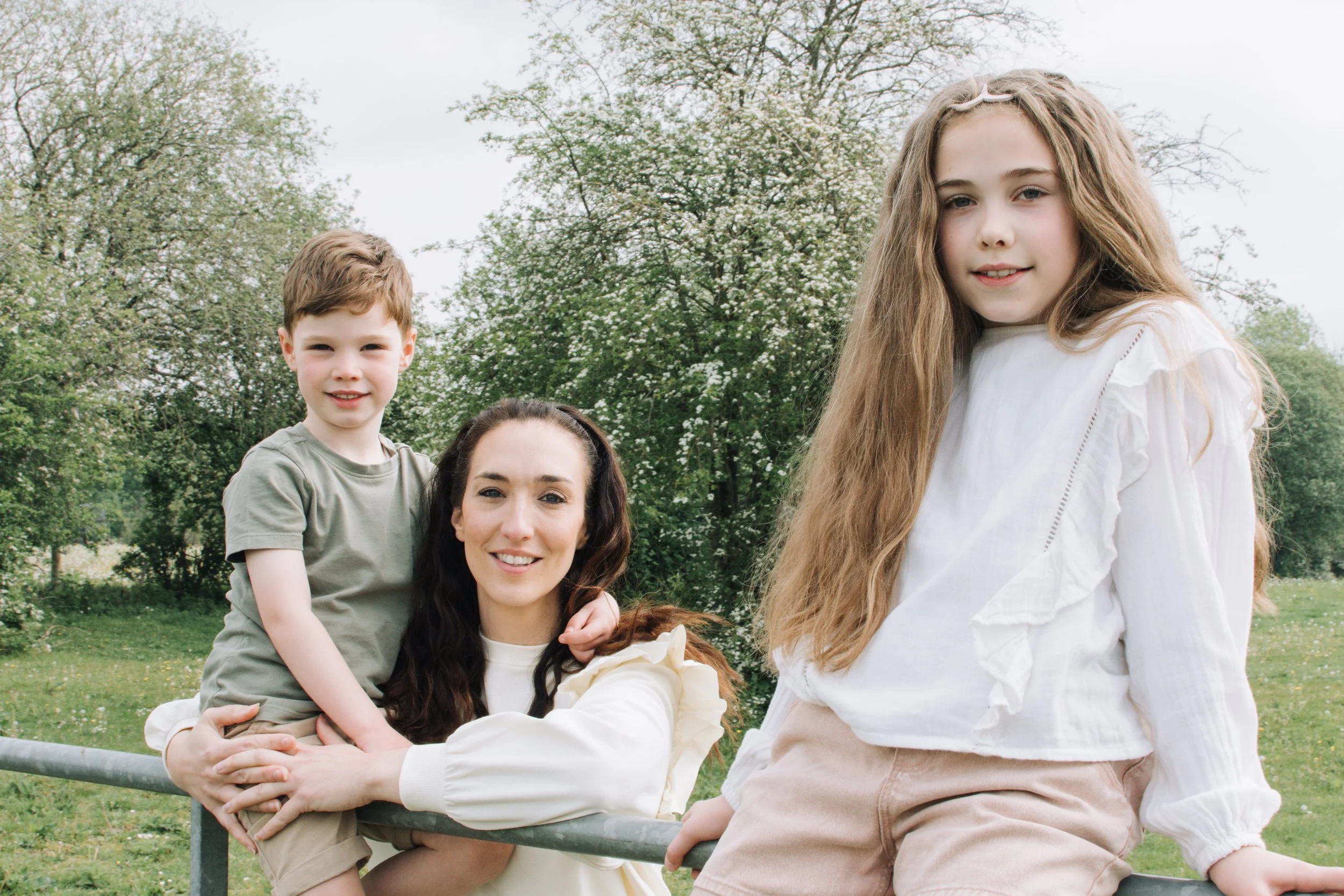 A woman and three children outdoors in a park with green trees in the background, smiling at the camera.