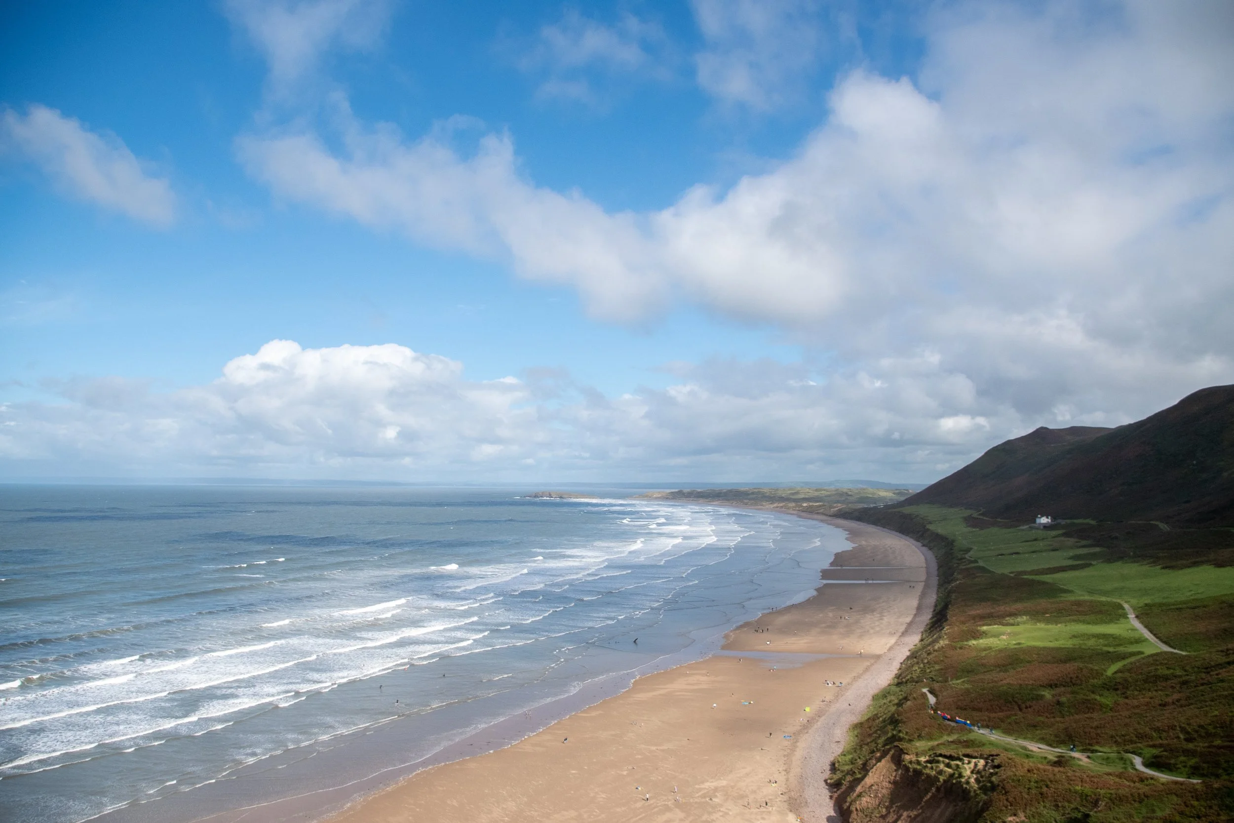 A scenic coastal view with a sandy beach, ocean waves, and green hills in the background under a partly cloudy sky.