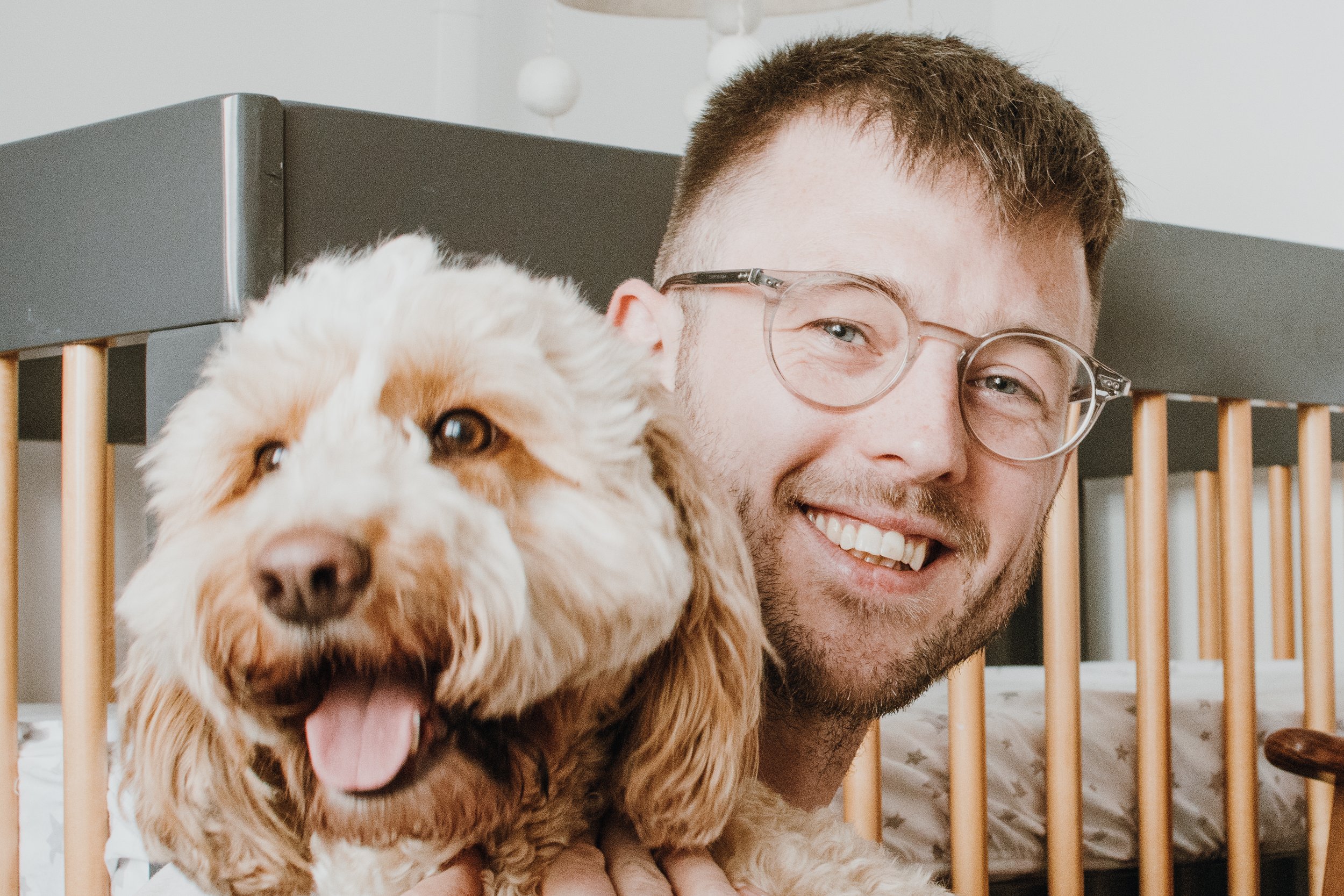 A smiling man with short hair and glasses next to a fluffy, light-colored dog with a pink tongue hanging out, inside a home with a wooden railing and white walls.